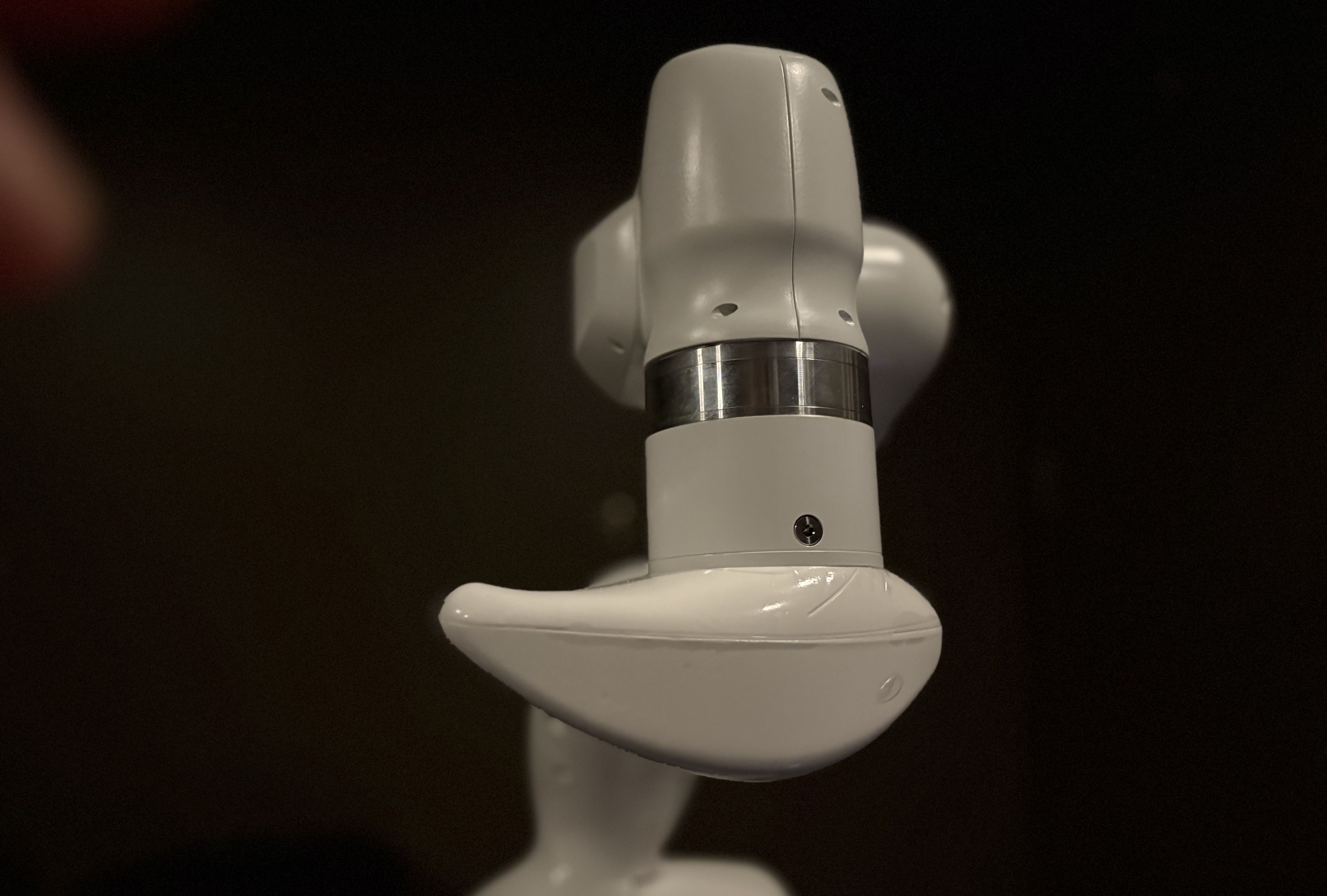 Close-up of a white robotic arm part with a curved grip and a metallic ring detail, set against a dark background.