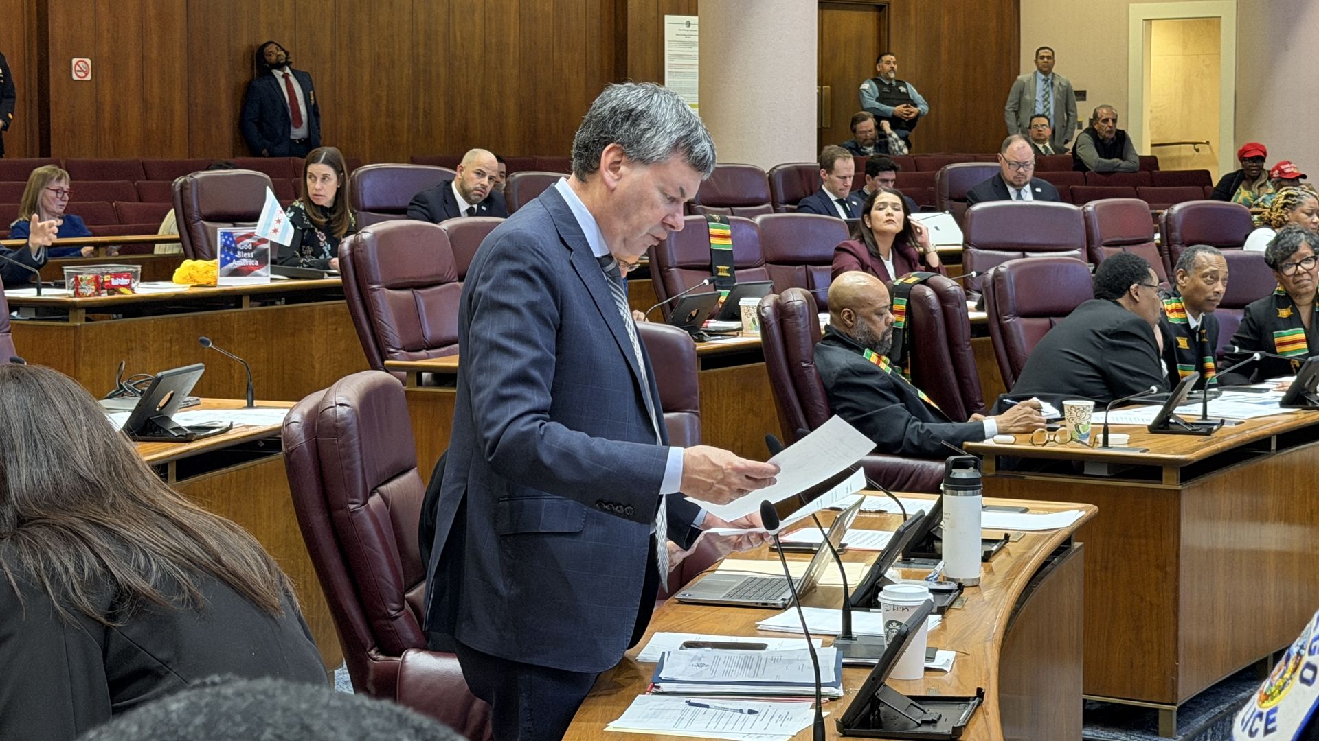 Inside a formal council chamber, a man in a suit reads documents at a desk while others sit in burgundy chairs. Wood-paneled walls, a central column, and security guards in the background.