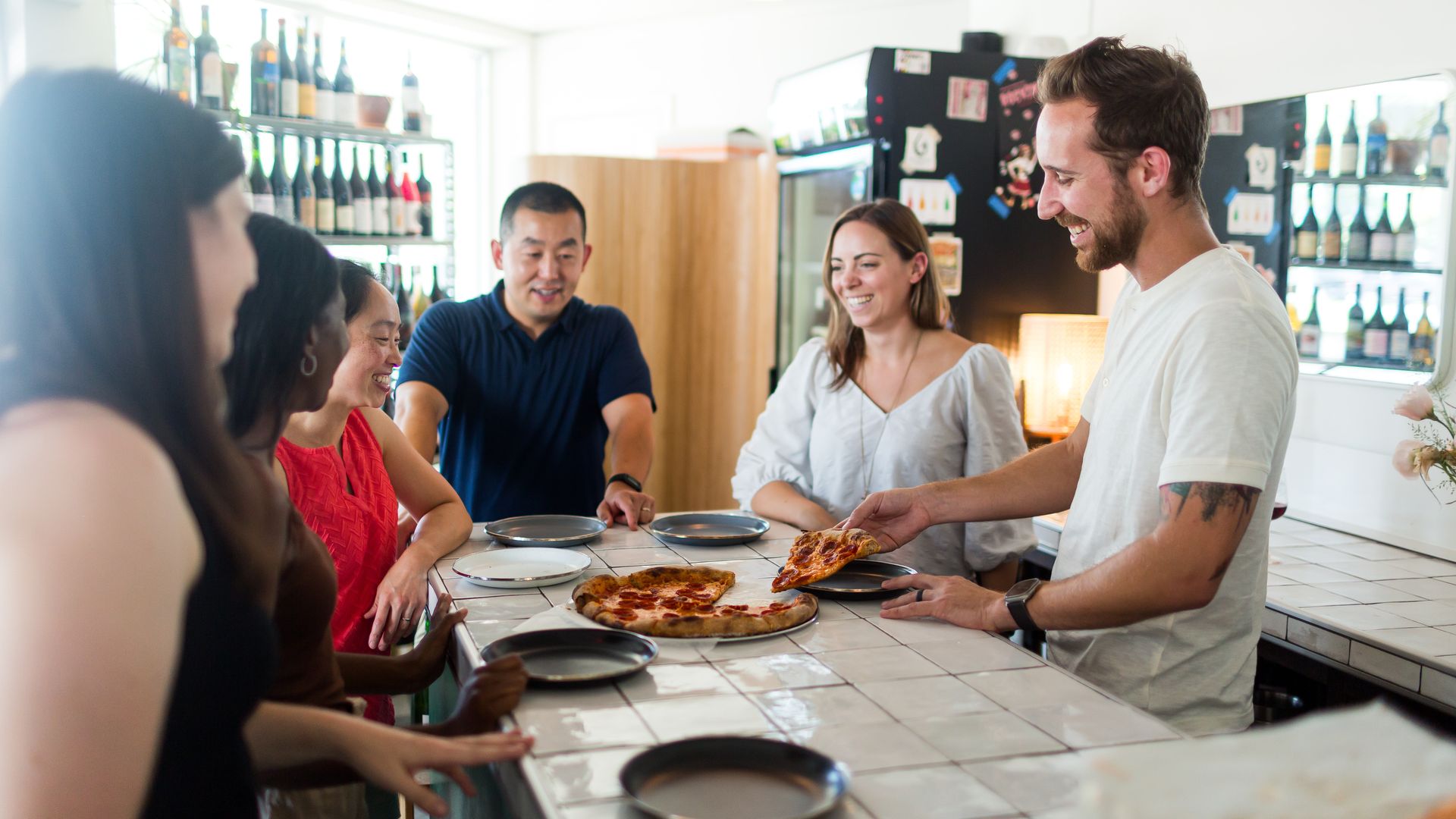 A group of people huddled around a pepperoni pizza with a background of wine bottles.