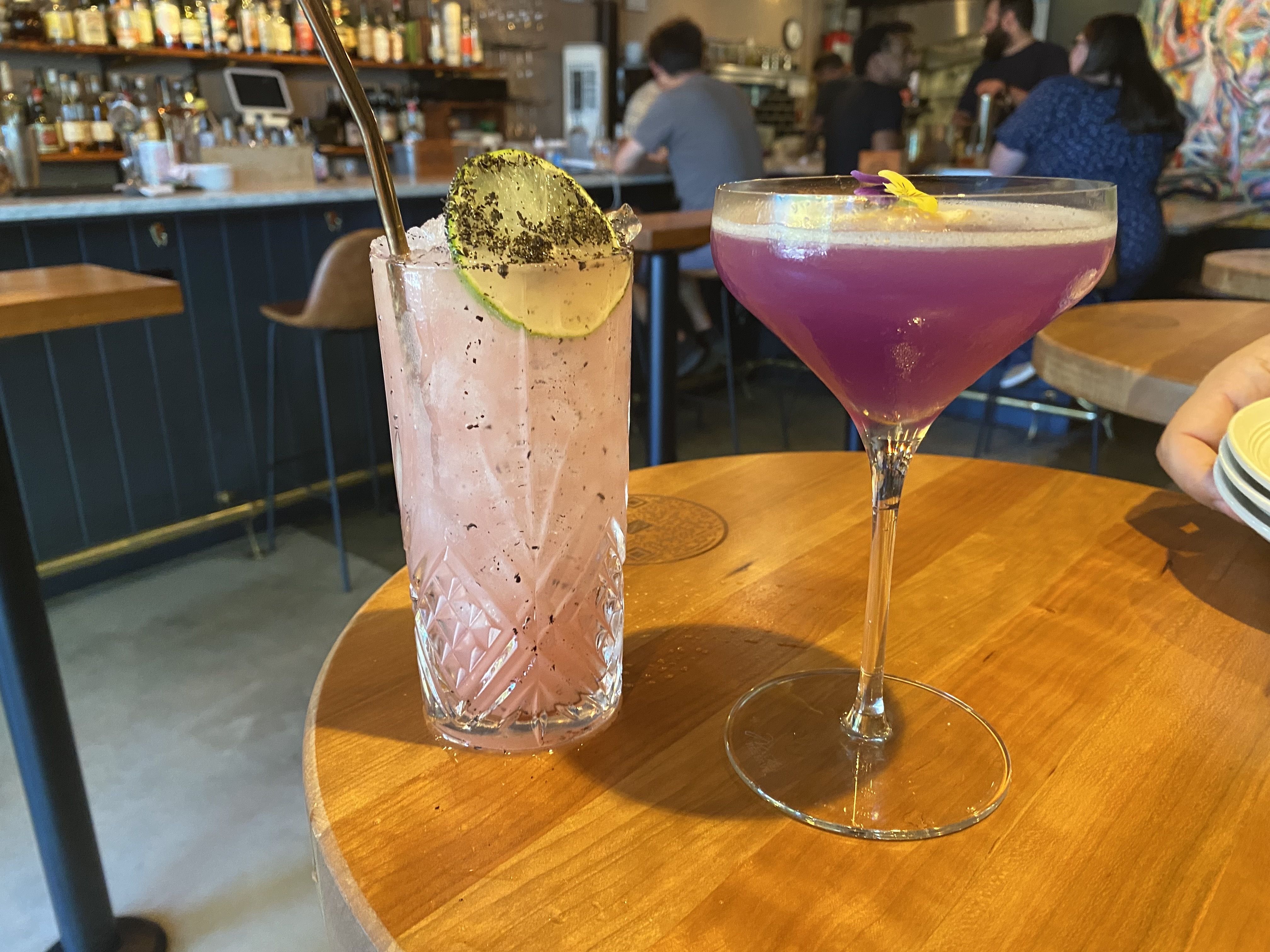 Two cocktails on a wooden table at a busy bar: a tall pink drink with a lime wheel and black specks, and a purple margarita in a coupe with a yellow flower; blurred patrons in back.