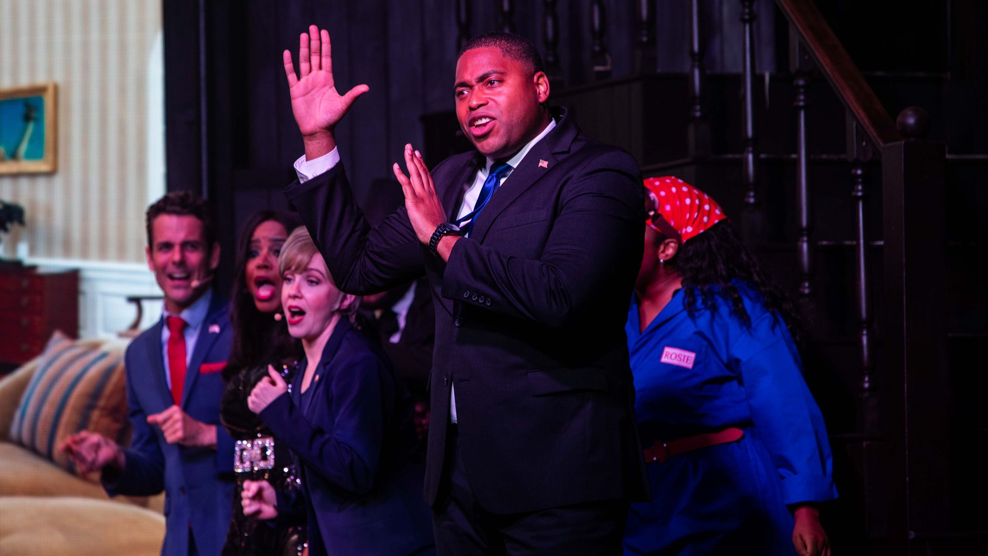 Man in suit with his hands in the air in front of people who are singing.