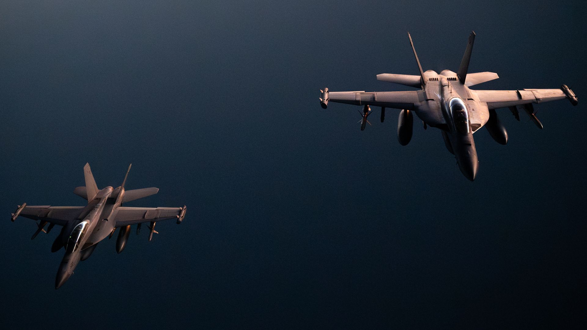 Two U.S. military fighter jets fly over open water in the Middle East. They are backlit by the sun.