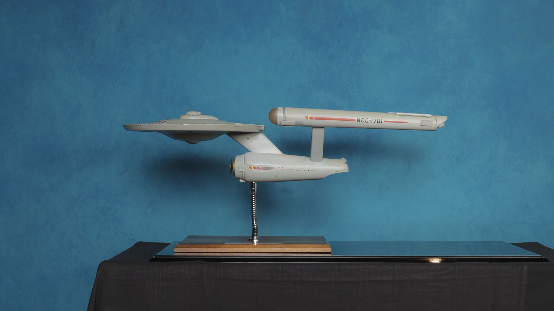 Scale model of the Starship Enterprise NCC-1701 on a metal stand, featuring a saucer-shaped hull and two warp nacelles, set against a blue textured backdrop.