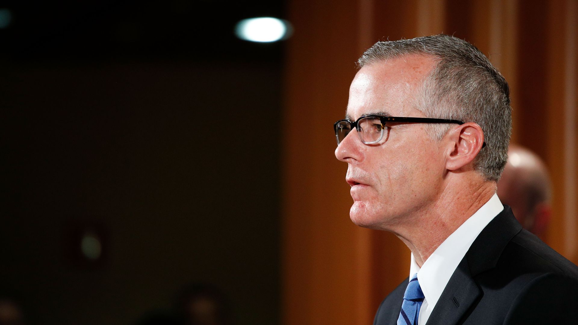 In this image, Andrew McCabe stands in a suit in front of a red curtain, looking to the left. 