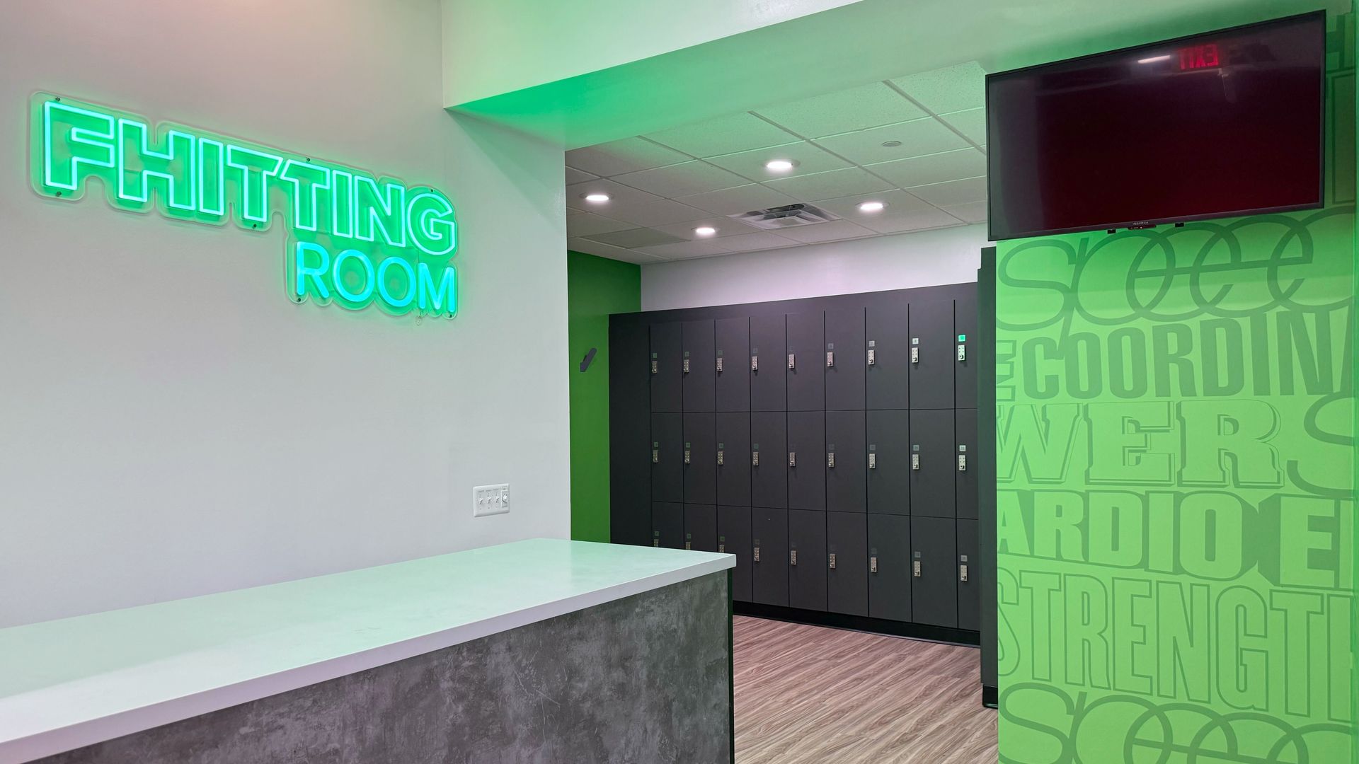 The interior of a gym with lockers and a neon sign reading "Fhitting Room." 