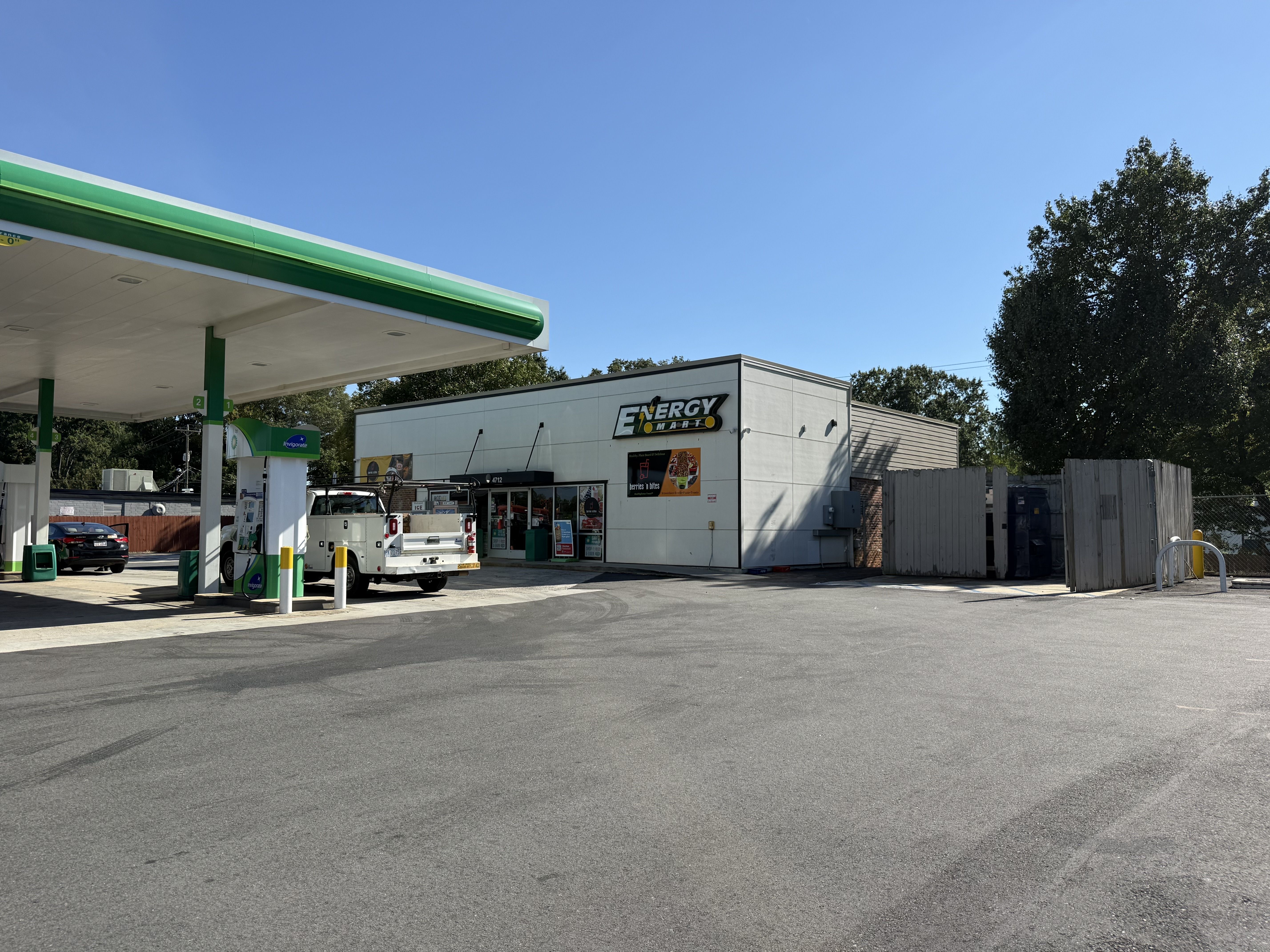 Wide gas station under a clear blue sky with a green-and-white canopy, fueling pumps, a white pickup, and a gray building labeled "Energy Mart"; trees line the right side.