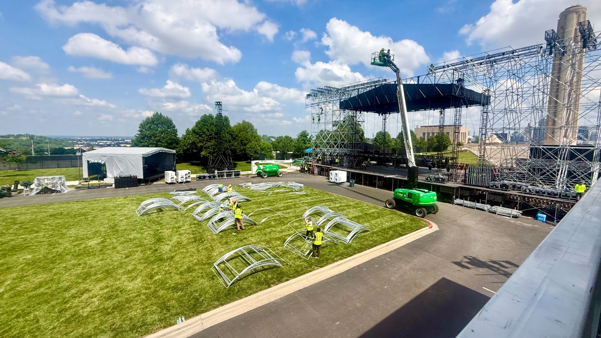 Outdoor concert stage under metal scaffolding with a crew in yellow vests assembling arches on a grass lot; a white tent, equipment, green lift, and a distant smokestack against a blue sky.
