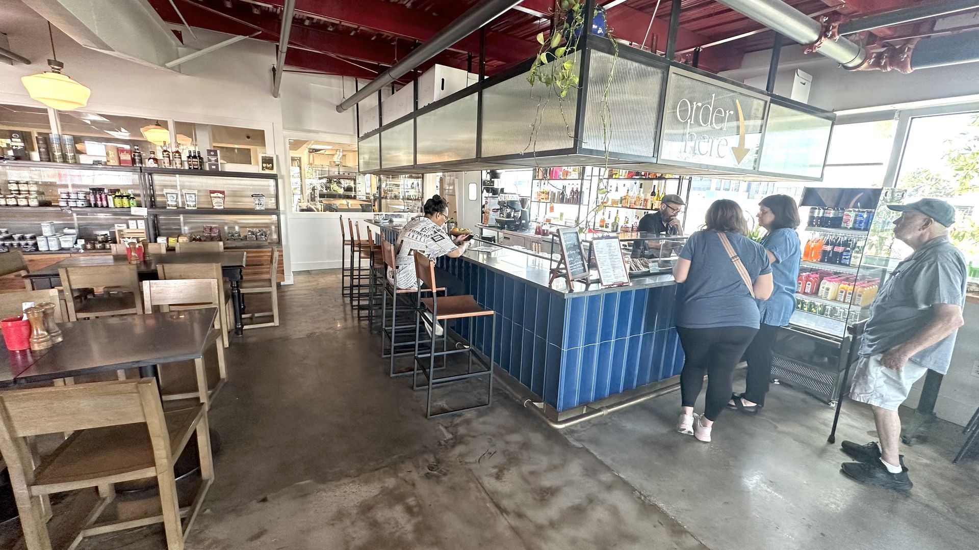 Interior of a cafe with wooden tables and chairs, a blue-tiled counter where one person sits, and three people plus a man with a cane standing near a sign saying "order here".