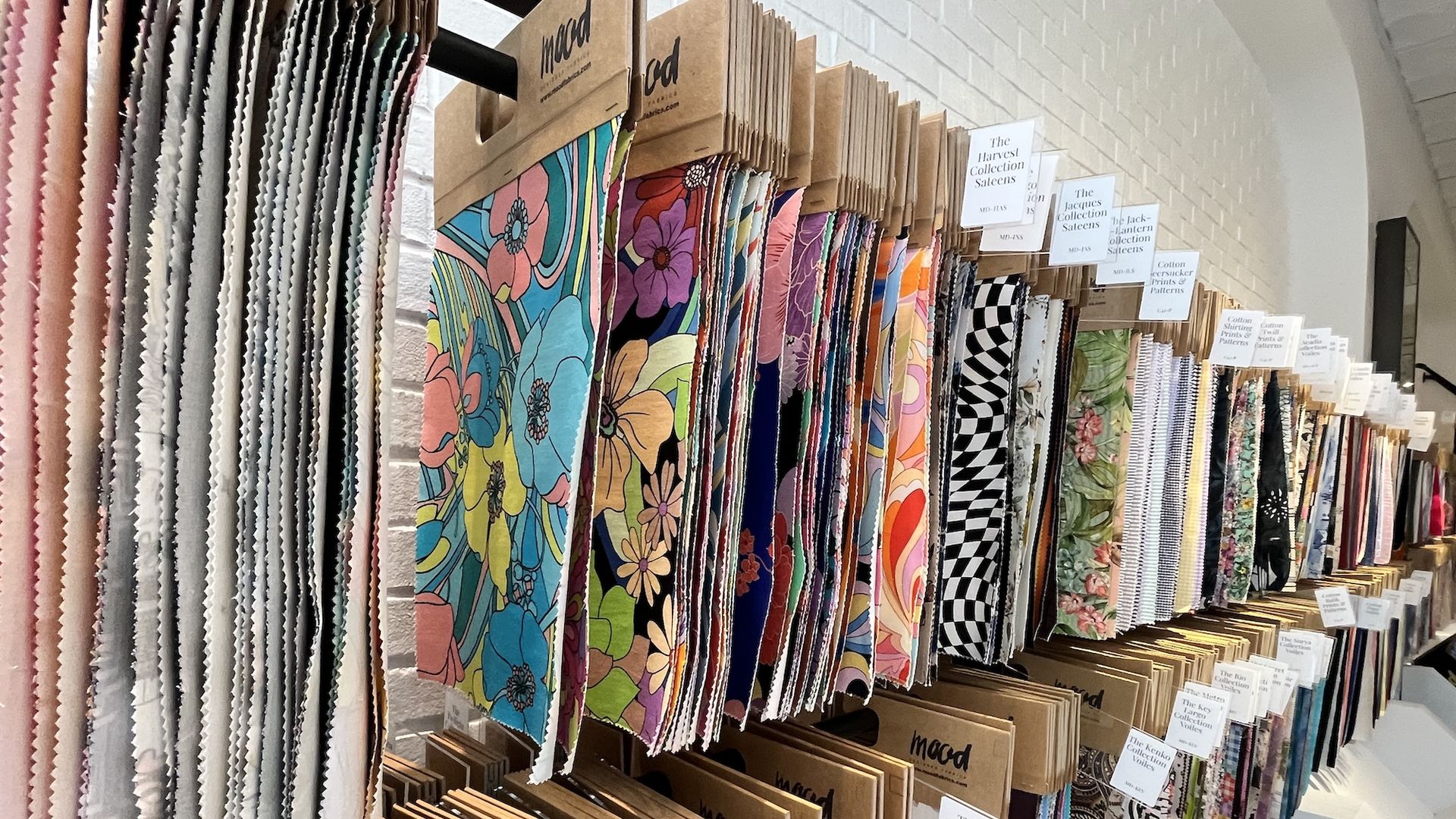 Row of fabric swatches on a display rack in a store, ranging from pastel pinks and neutrals to bold floral prints and black-and-white checks, with mood-brand tags above.