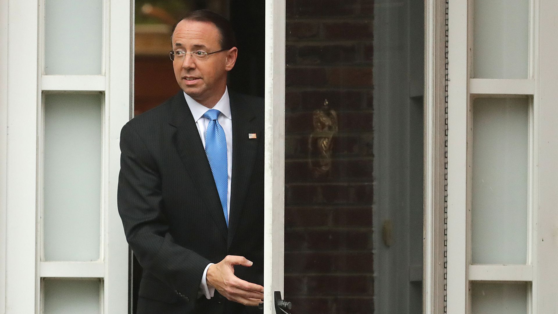 Rod Rosenstein in a suit and light blue tie pushing open a glass door as he steps outside of a building. 