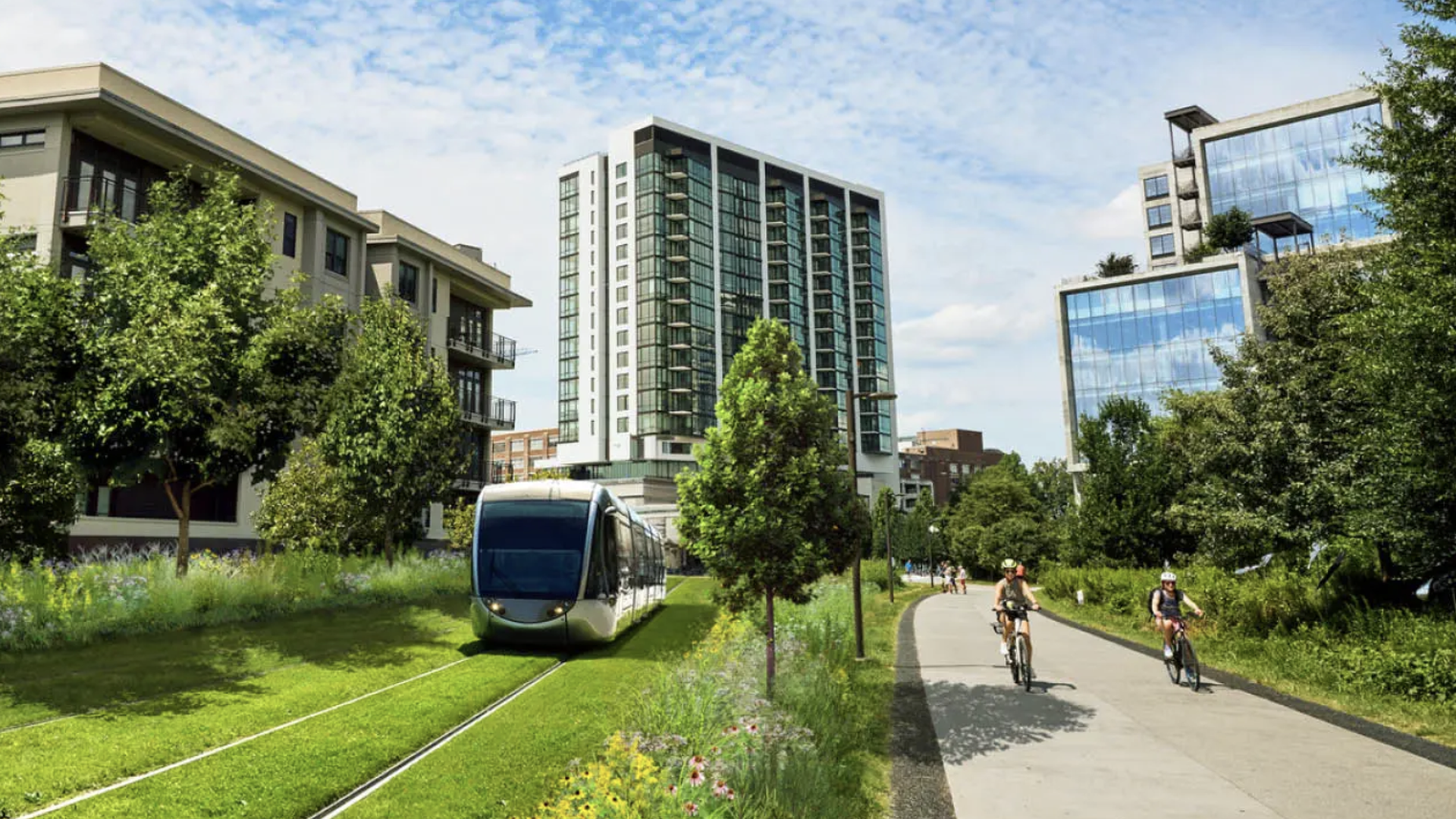 Modern light rail train on green tracks beside a path with cyclists, surrounded by trees, flowers, and modern buildings under a blue sky with scattered clouds.