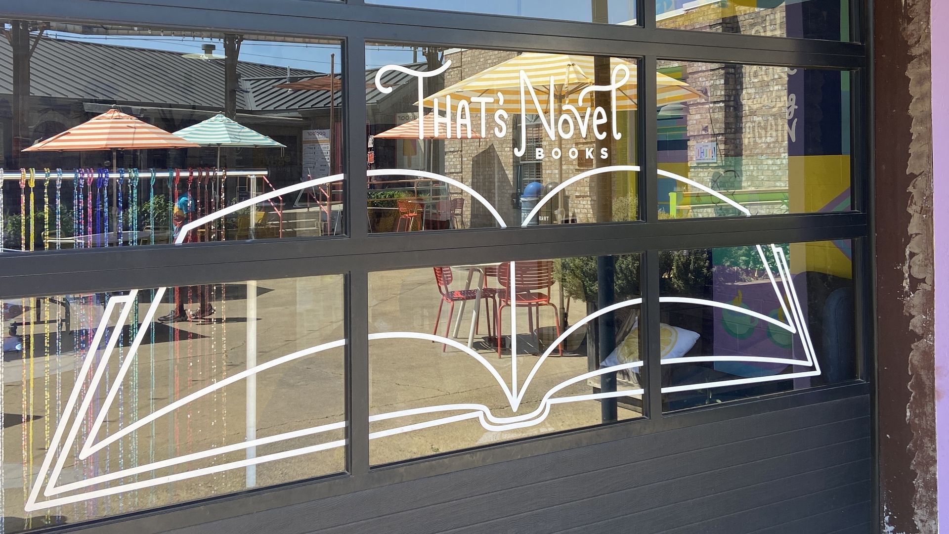 Glass door of That’s Novel Books with a white open book design and text, colorful umbrellas and chairs reflected, brick wall painted pink with yellow and green shapes above.