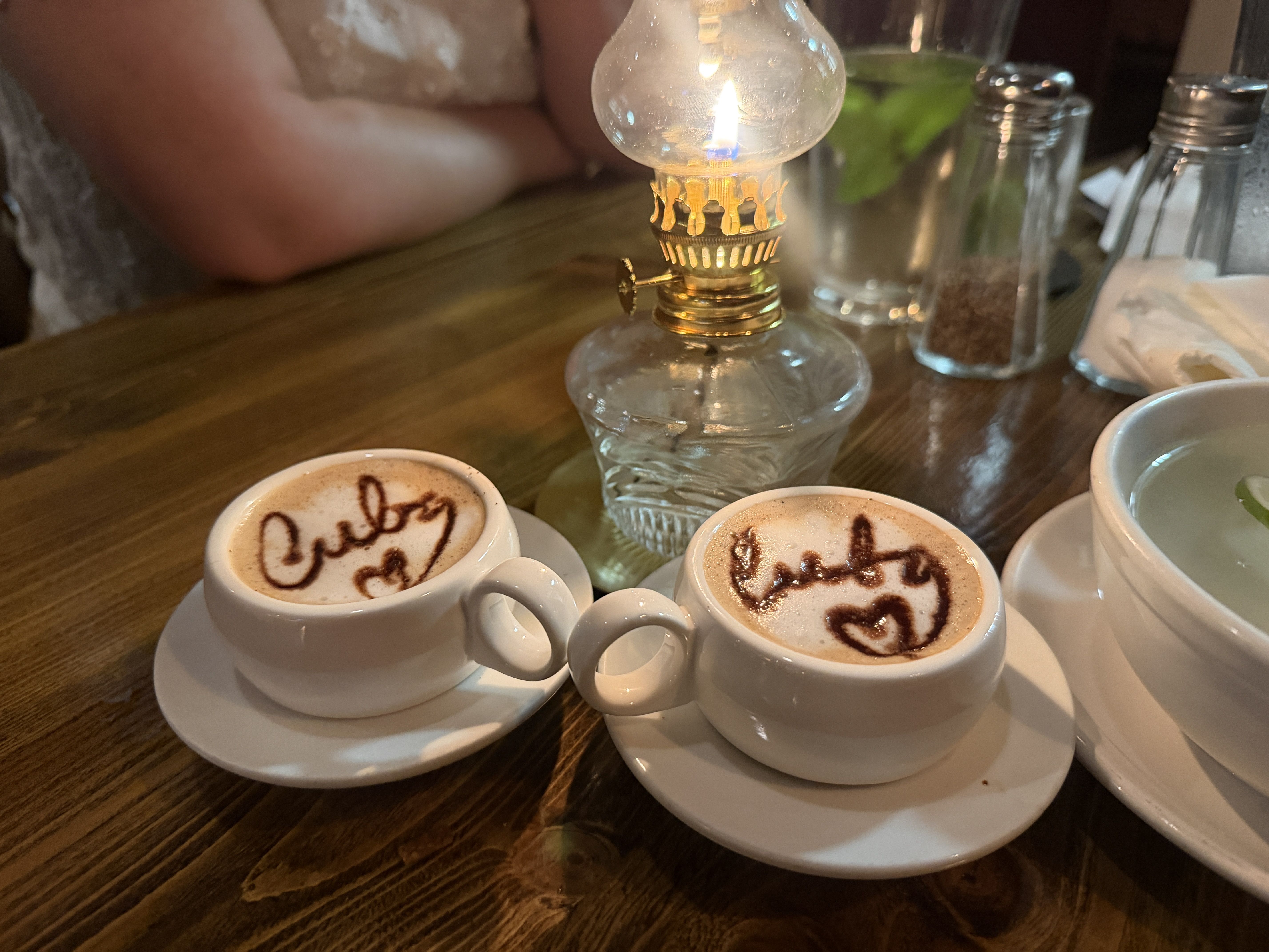 Three white cups of cappuccino on saucers with cocoa-script foam around a vintage glass oil lamp on a wooden table; blurred background shows drinks, shakers, and a napkin.