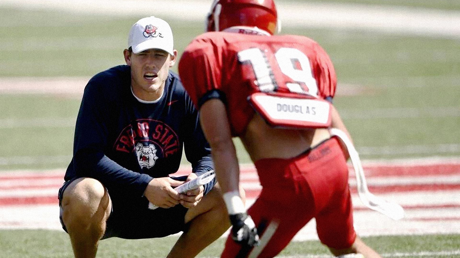 KIrby Moore crouches wearing a long sleeved Fresno State T-shirt and baseball cap, holding papers while looking at a red-jerseyed football player who has his back to the camera, on a football field.