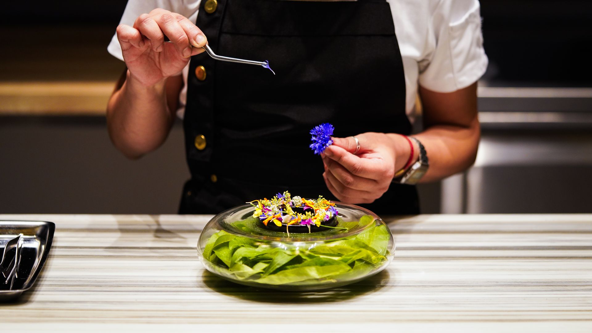 A chef uses tweezers to place small, purple flower petals on an intricate dessert.