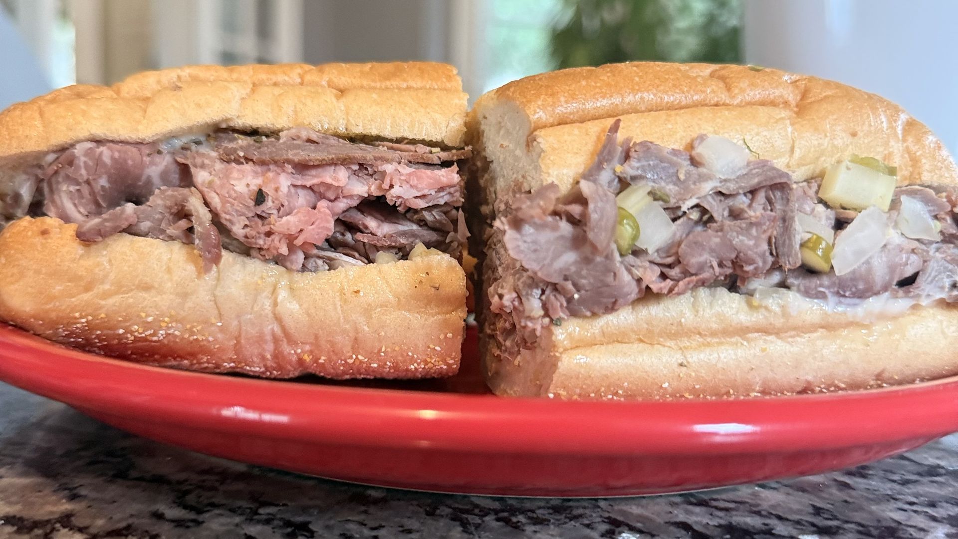 Two beef sandwiches on a red plate sit on a dark granite countertop. Left half shows sliced roast beef; right half has chunky meat with onions and pickles. Blurred indoor background.