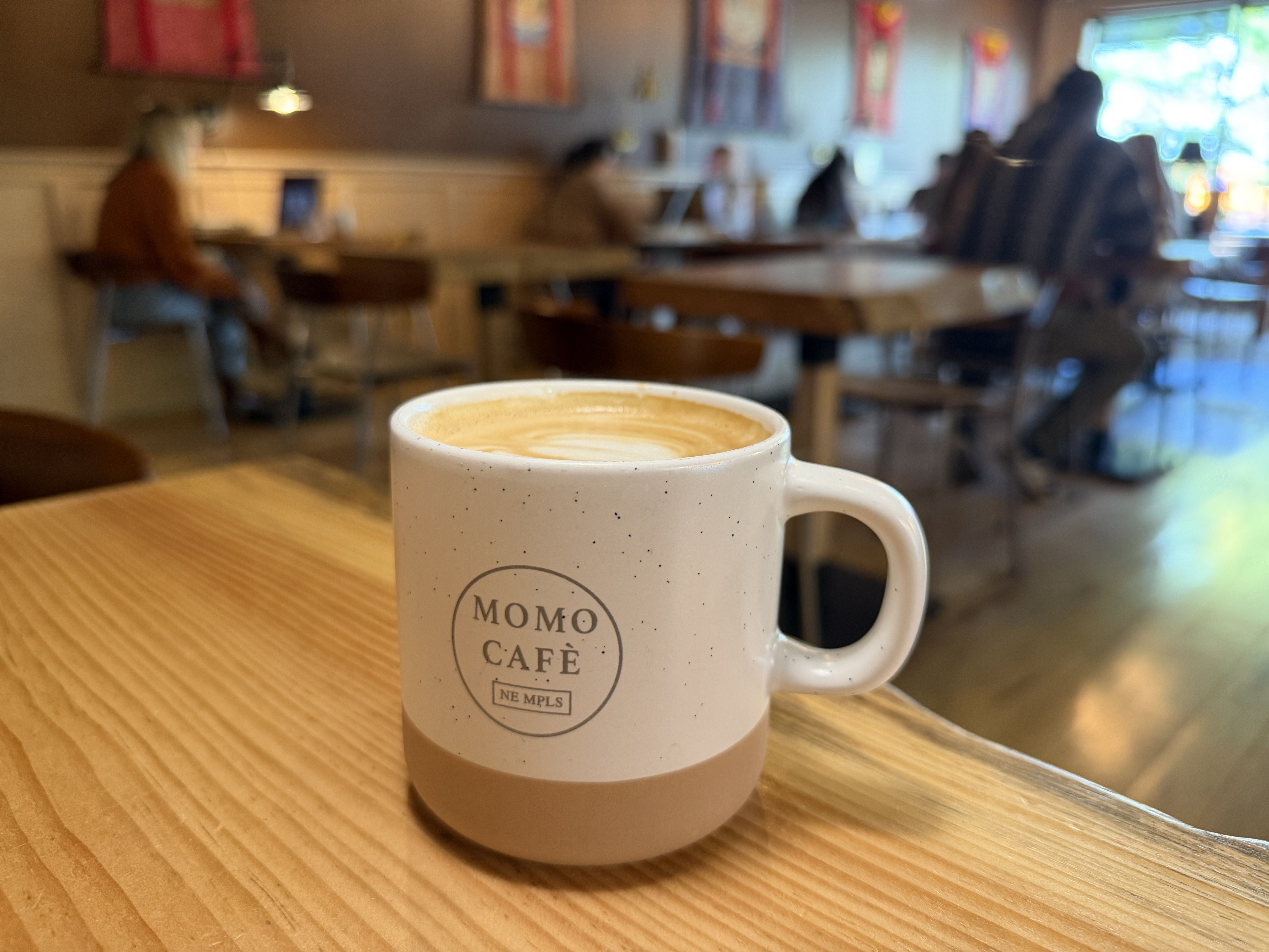 White speckled mug with the MOMO CAFE logo sits on a wooden table, latte art on top; blurred cafe interior with patrons and a bright window in the background.