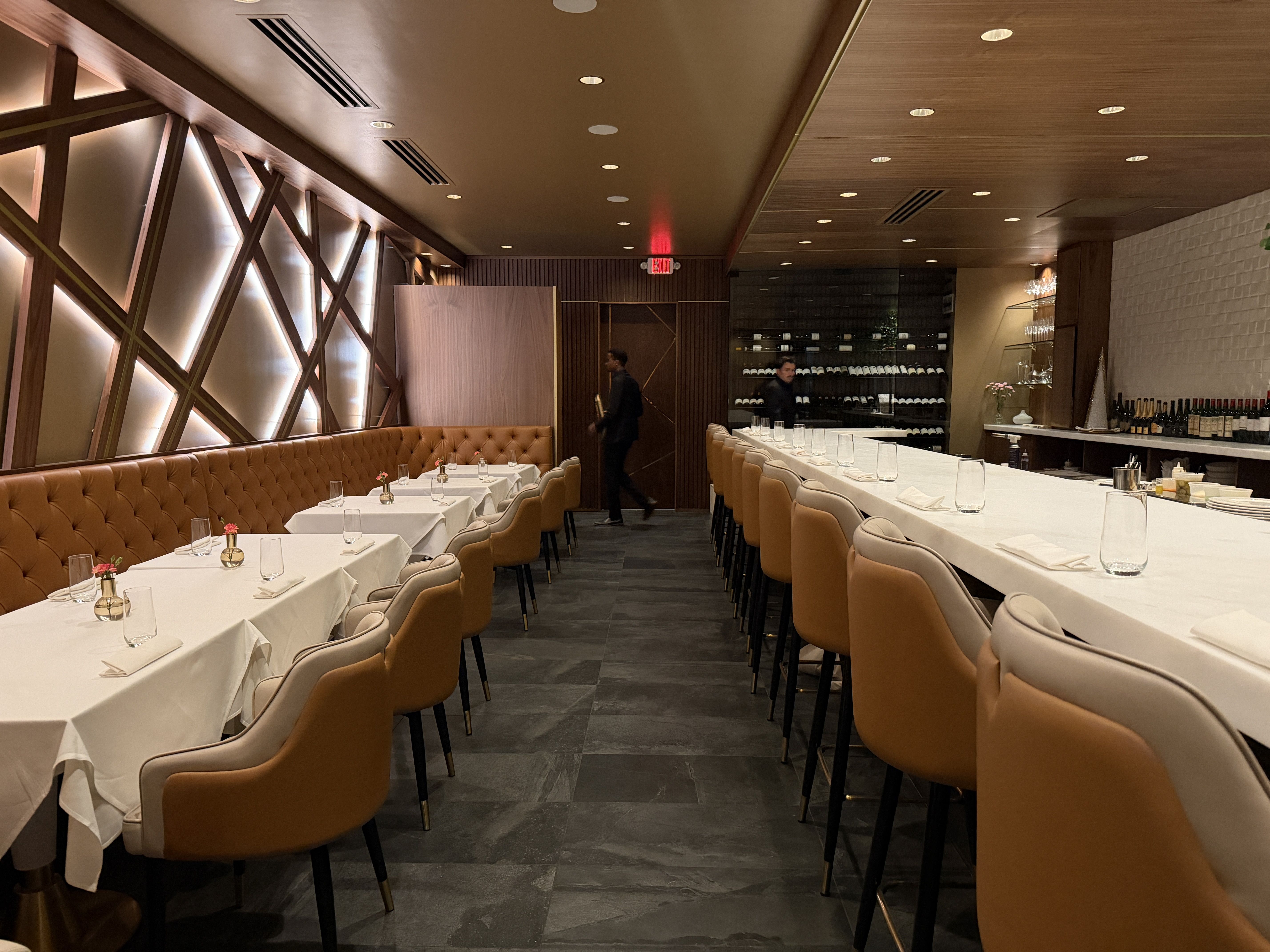 Interior of a modern restaurant with tan leather chairs, white tablecloths, small flower vases on tables, geometric wooden wall design, and wine bottles displayed behind the bar.