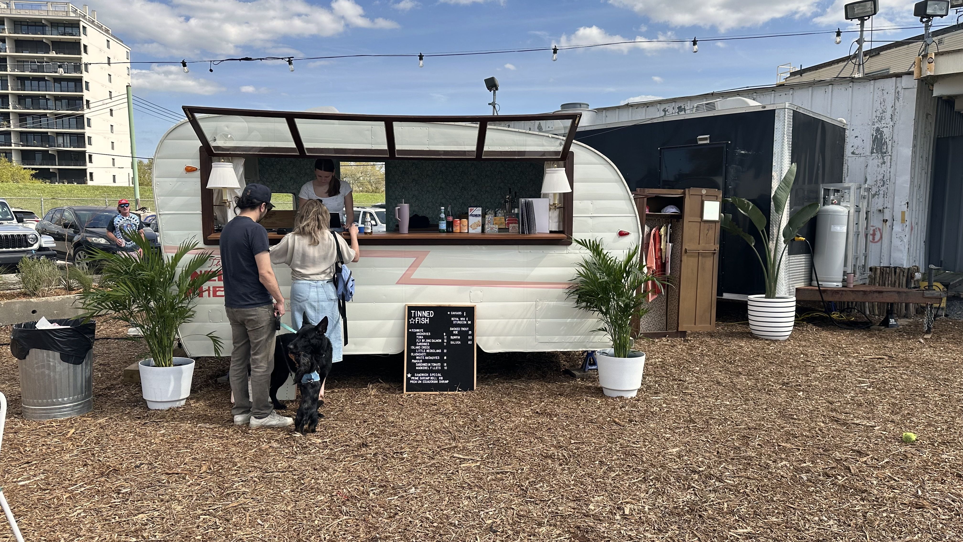 Two people order food at a hip-looking trailer-turned-food truck. One of the people holds a leashed dog while another, smaller dog, plays with the larger one.