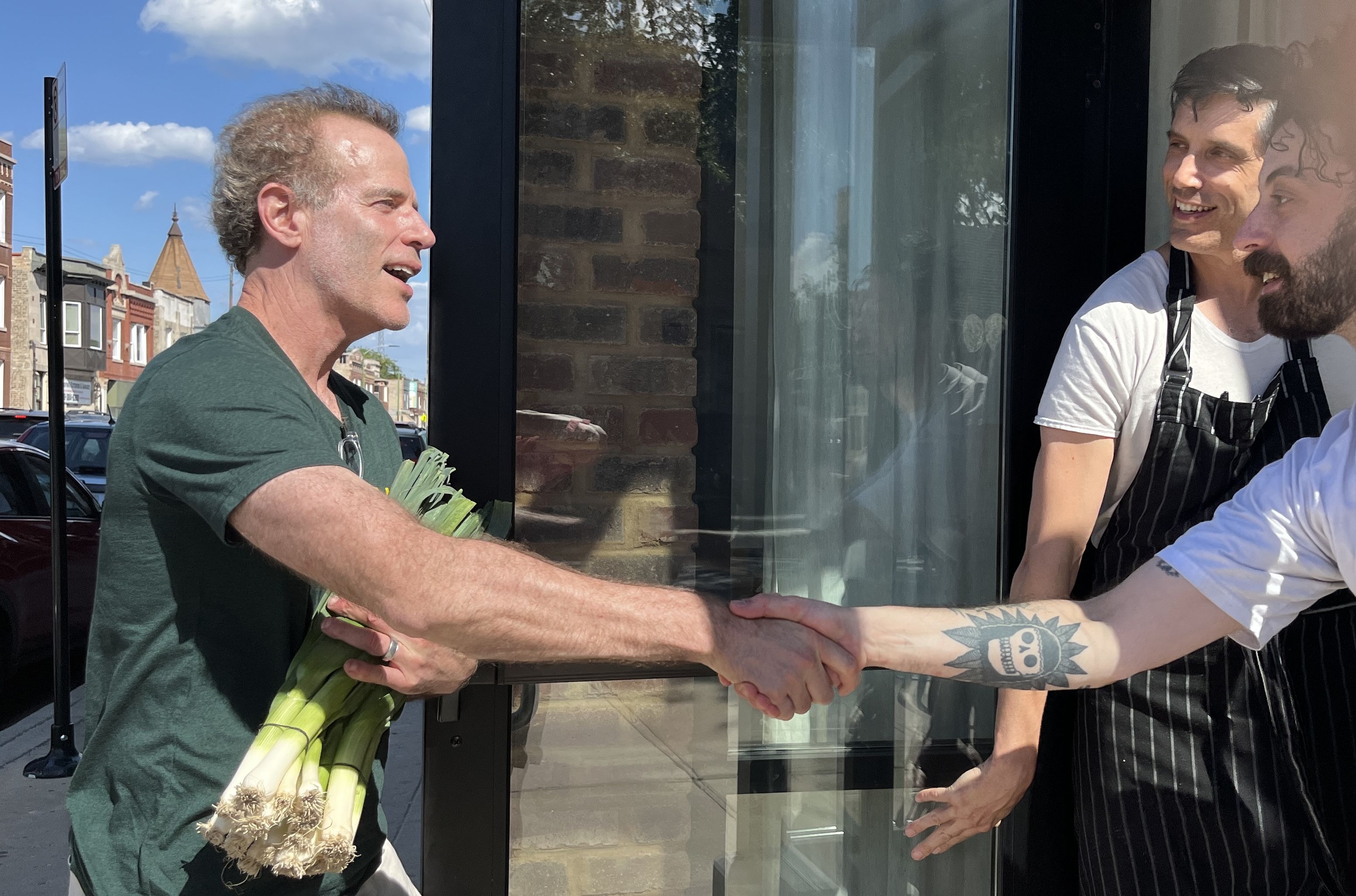 Man in green shirt holding leeks shakes hands with tattooed man in white shirt and striped apron outside brick building with blue sky and street in background.