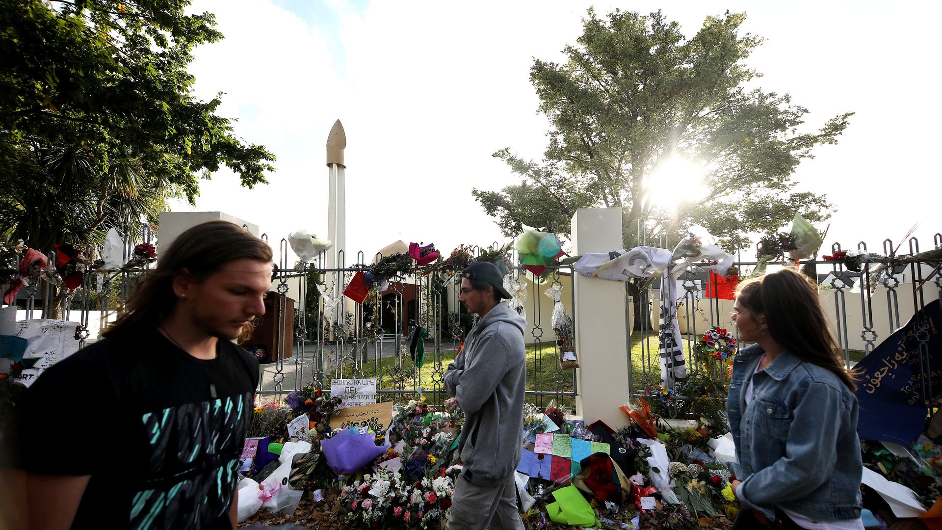 In this image, people walk by flowers and memorials laid out near a mosque fence.