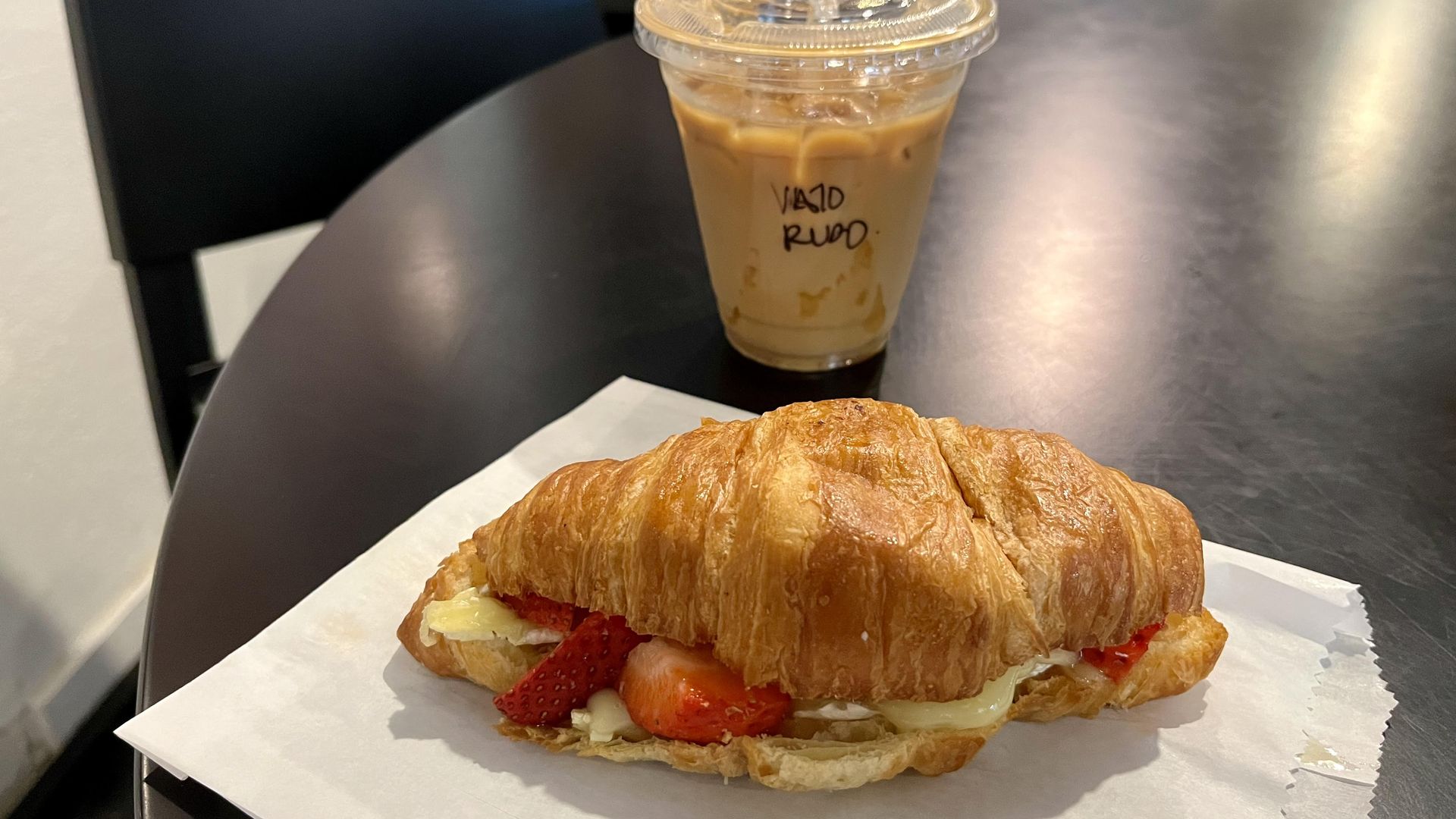 A warm brie, berries and honey croissant next to a coffee in a to-go cup that says "Vato Rudo" on it.