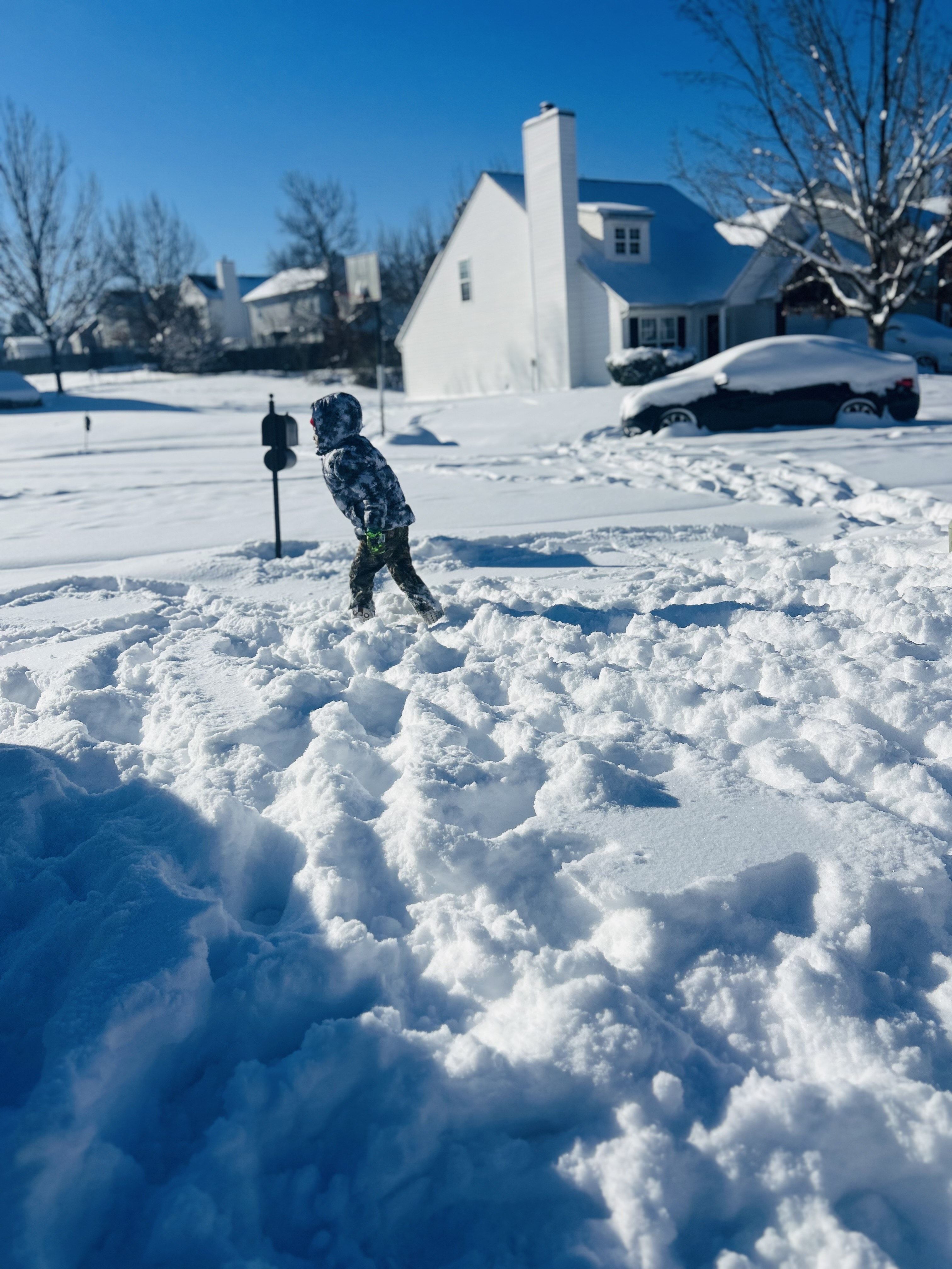 Child in a black and white coat walking through deep snow on a sunny winter day, with snow-covered cars and houses under a clear blue sky in the background.