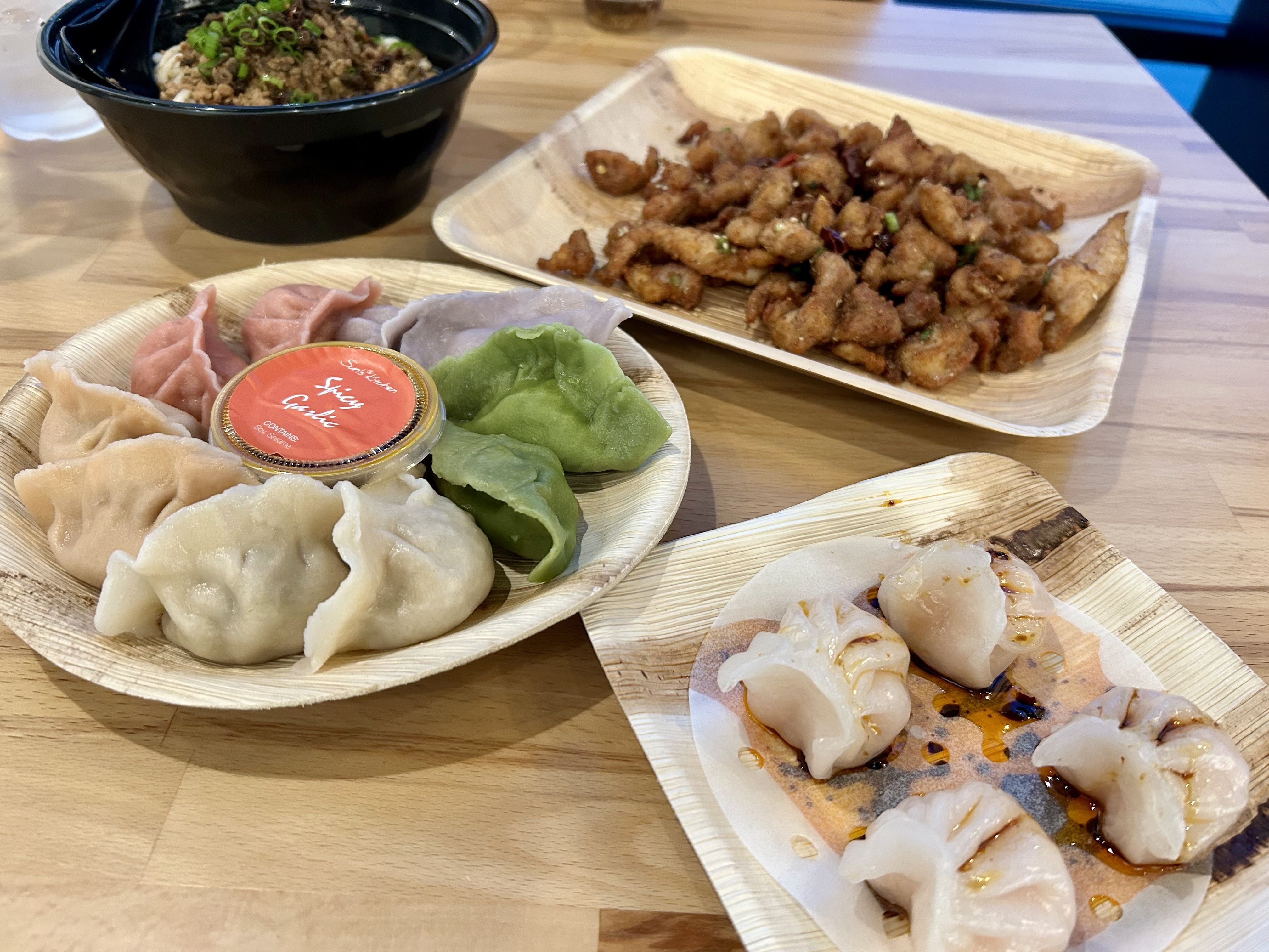 A variety of Asian dishes on a wooden table. In the foreground, several colorful dumplings are arranged on a round, wooden plate, accompanied by a small container labeled "Spicy Garlic." Near them, on another plate, are lightly seared dumplings with a drizzle of sauce. 