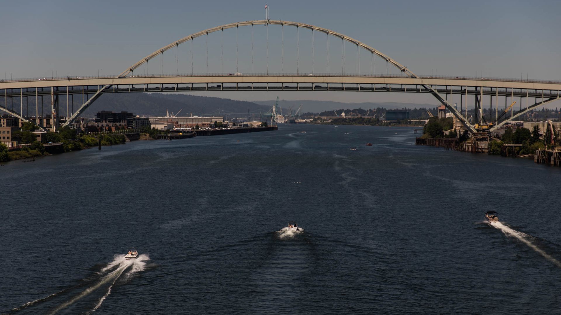 Wide river with a large steel arch bridge overhead. Road deck supported by suspenders; city on the far bank, hills in the distance. Several boats speed across, leaving wakes.
