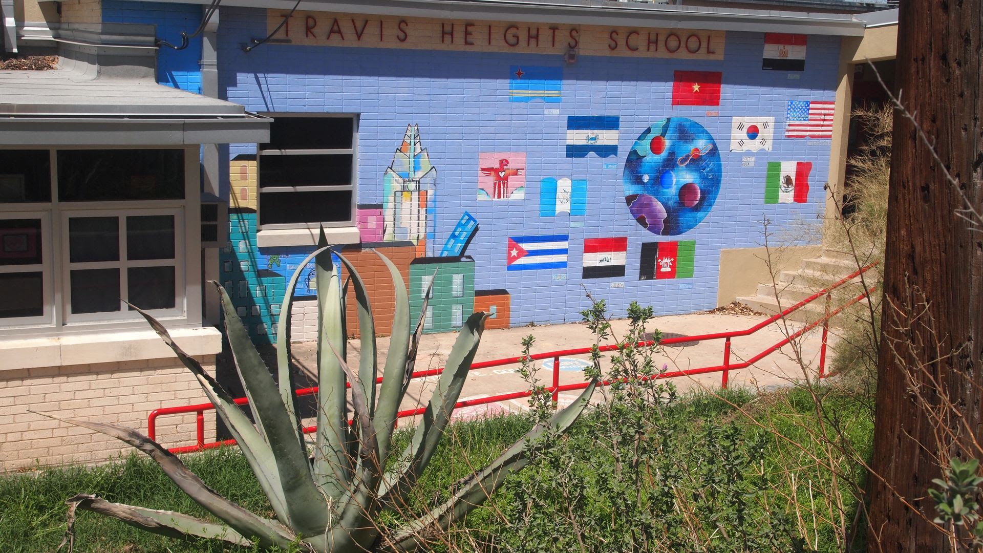 Blue brick wall mural at a school reading "TRAVIS HEIGHTS SCHOOL" with many flags and a globe; foreground shows a large agave plant and a red railing along a sidewalk.