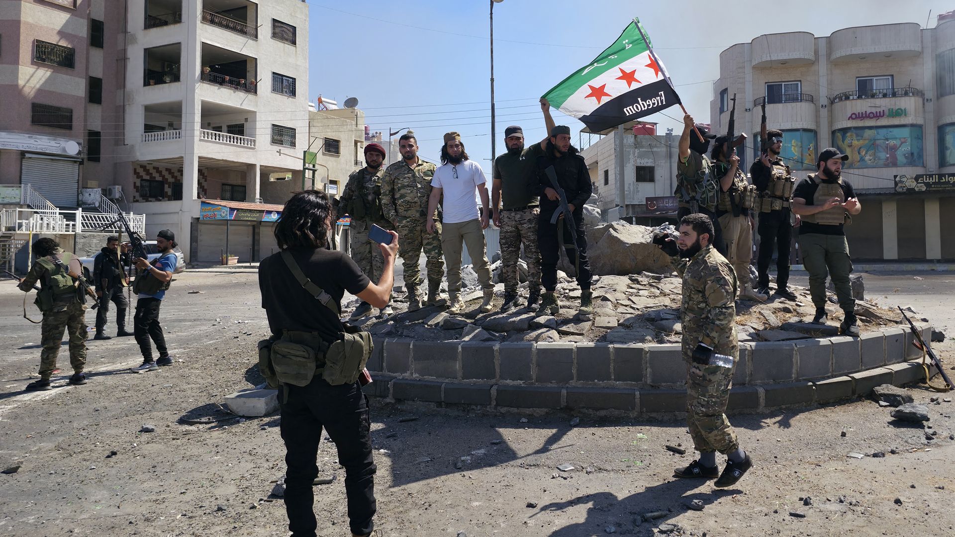 Group of armed men in military and civilian clothes gathered on a circular stone platform, one holding a green, white, and black flag with red stars and the word "Freedom" in an urban area.