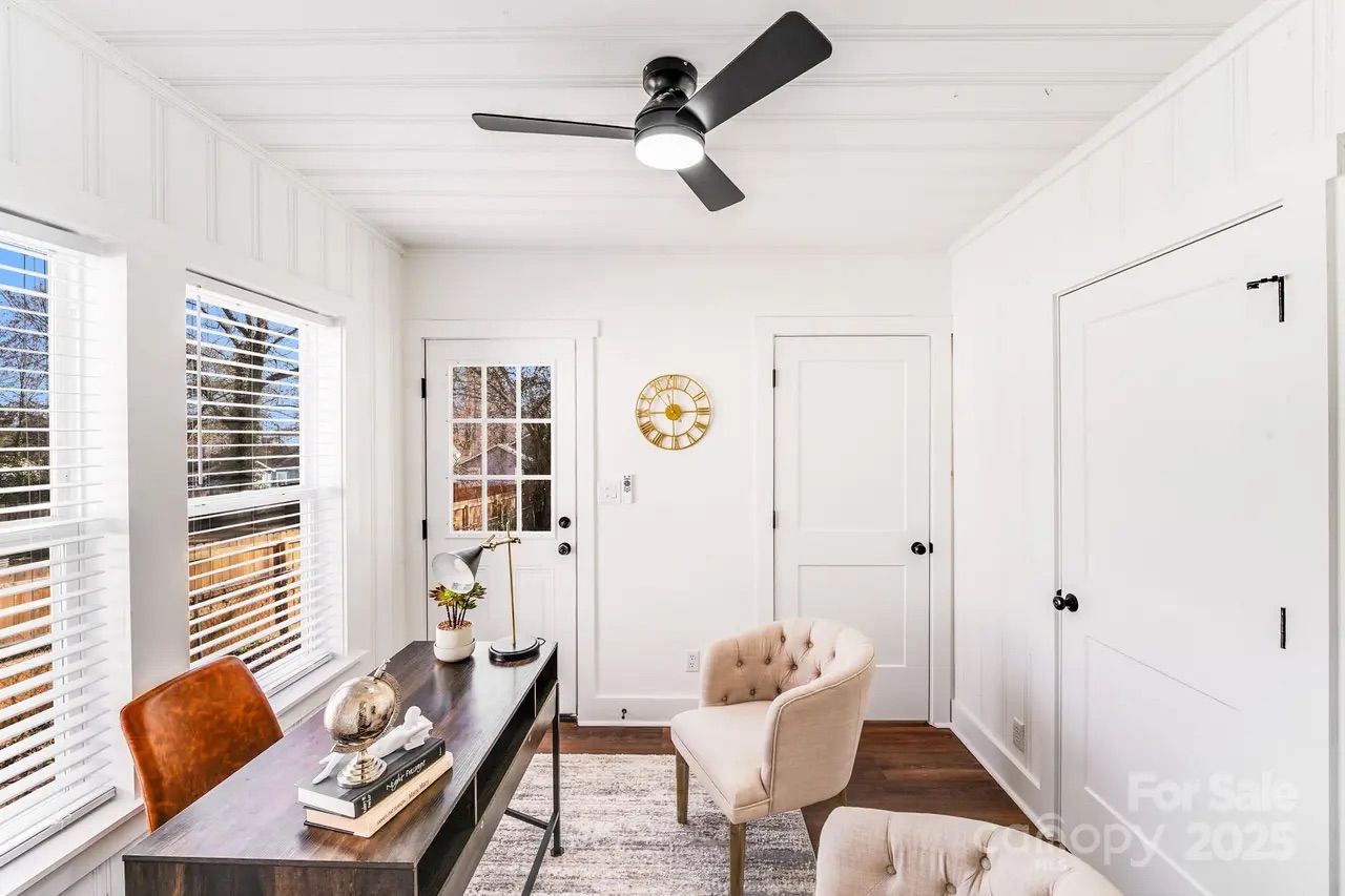 Bright white home office with dark wood desk, brown leather chair, beige armchairs, gold wall clock, black ceiling fan, large windows with white blinds, and wooden floor.
