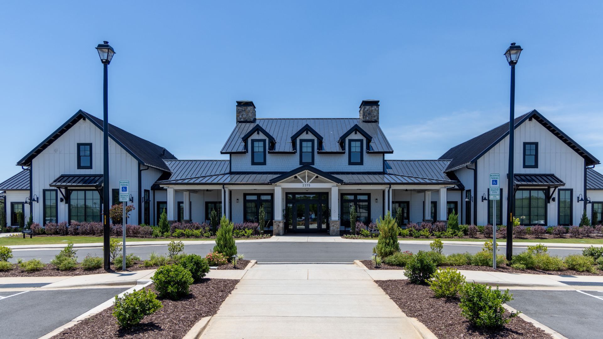 Front view of a white, modern farmhouse with dark blue trim and a metal roof. Central block shows three dormers and two chimneys; two tall lamps frame the entrance under a clear blue sky.