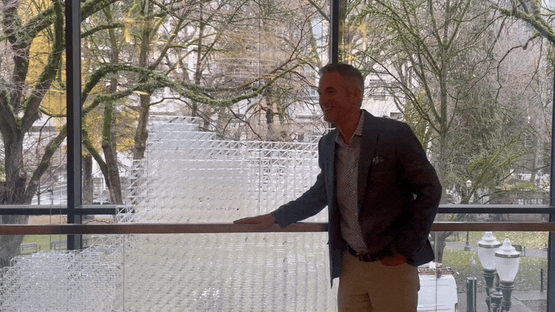 A moving image of a person standing near a window of a building and facing the camera then smiling.