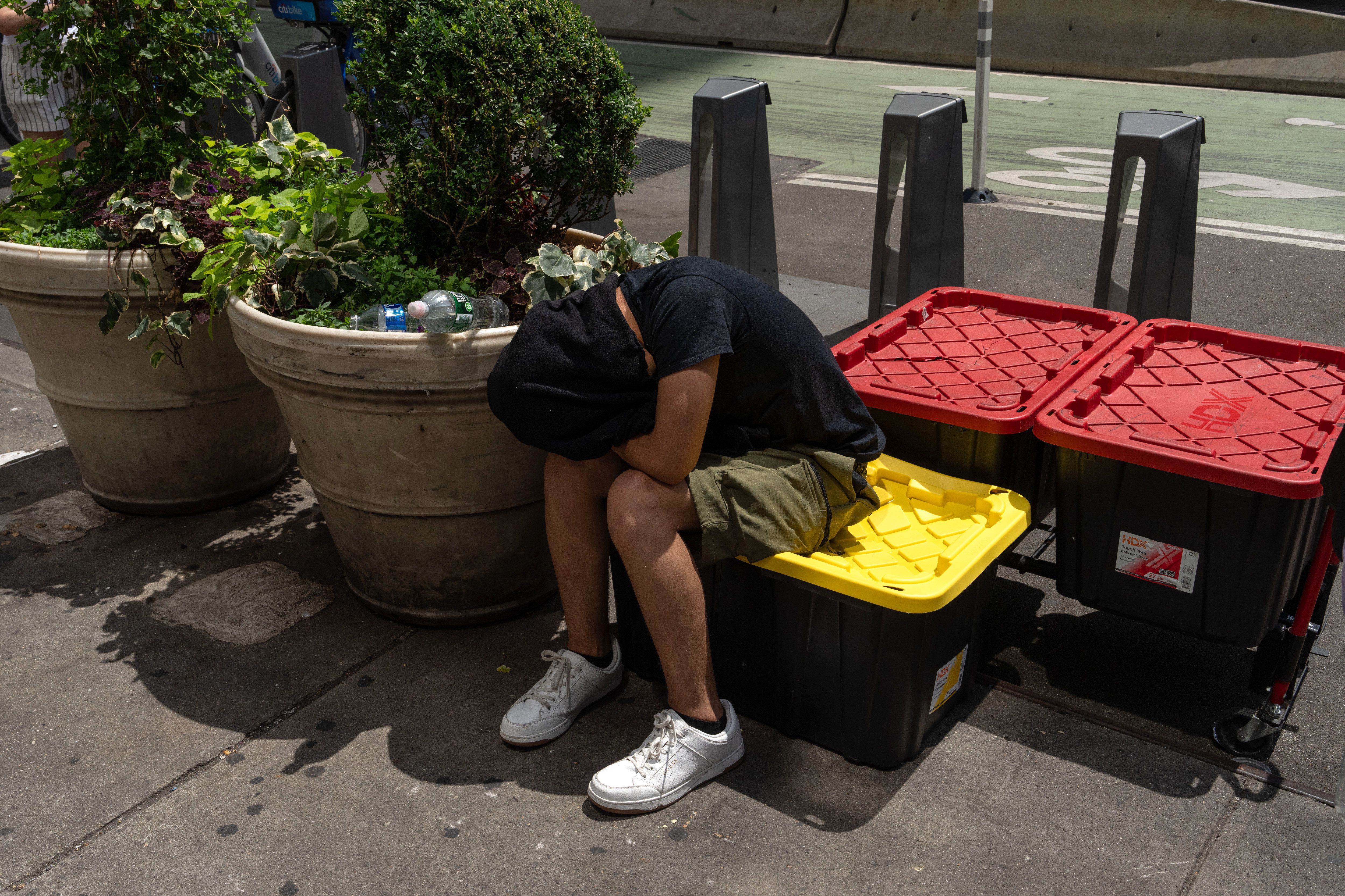A person leaning against a large flower pot on the sidewalk with their head covered to escape the heat. 