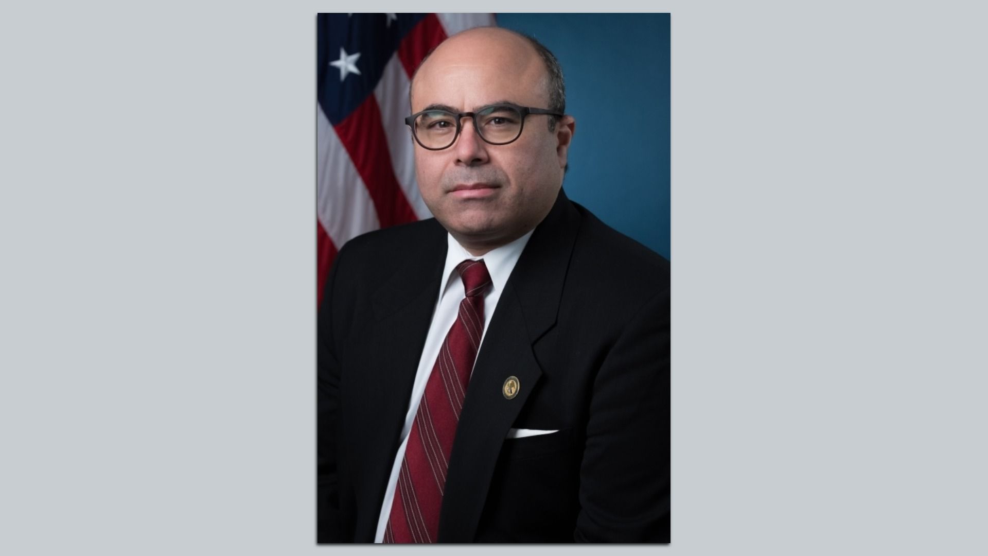 Formal portrait of a middle-aged man wearing a black suit, white shirt, red striped tie, and glasses, with an American flag and blue background behind him.