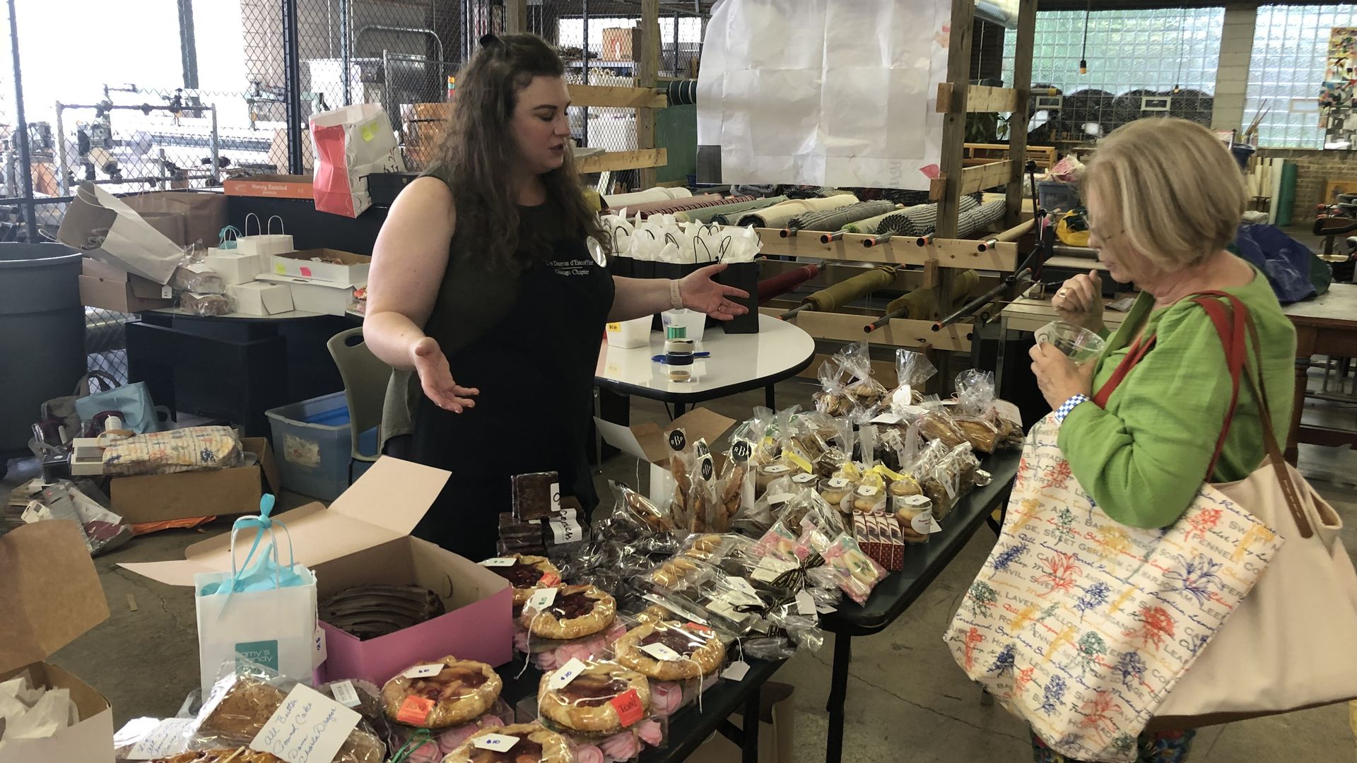 Photo of a worker at a bakery table talking to a customer.