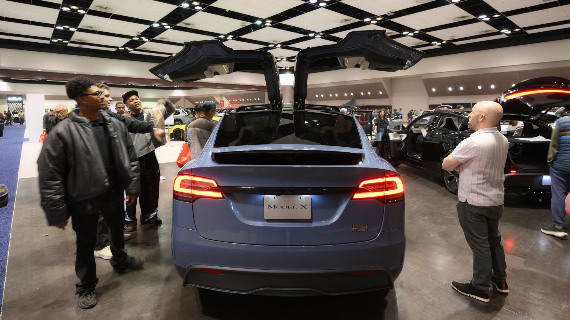 Blue Tesla Model X with open falcon-wing doors at an indoor car show; group of people nearby, bright overhead lights, other cars and attendees in the background.