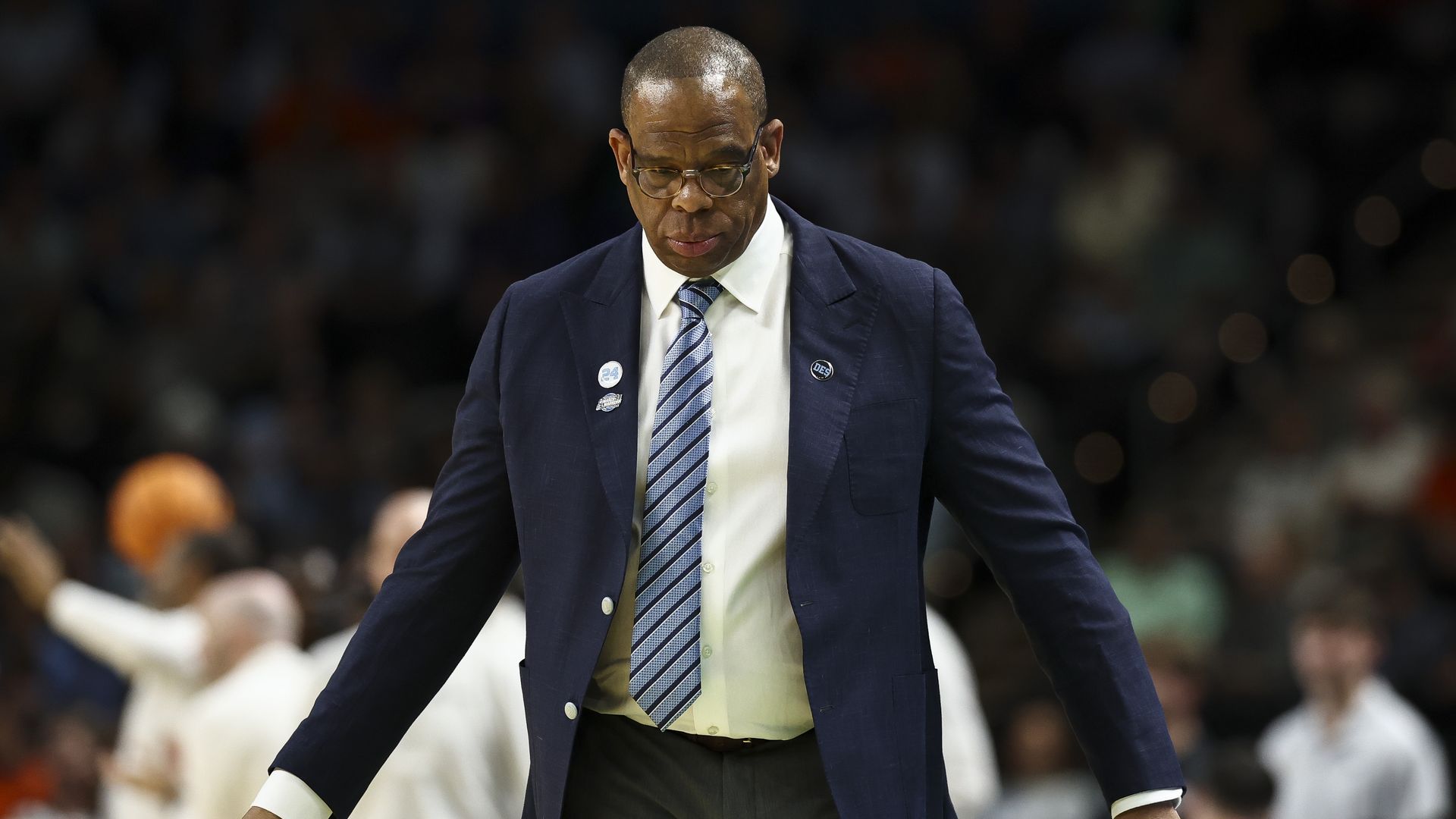 A man in a navy blazer, white shirt, and striped blue tie walks across a basketball court. He wears glasses and pins on his lapels; a blurred crowd fills the stands behind him.
