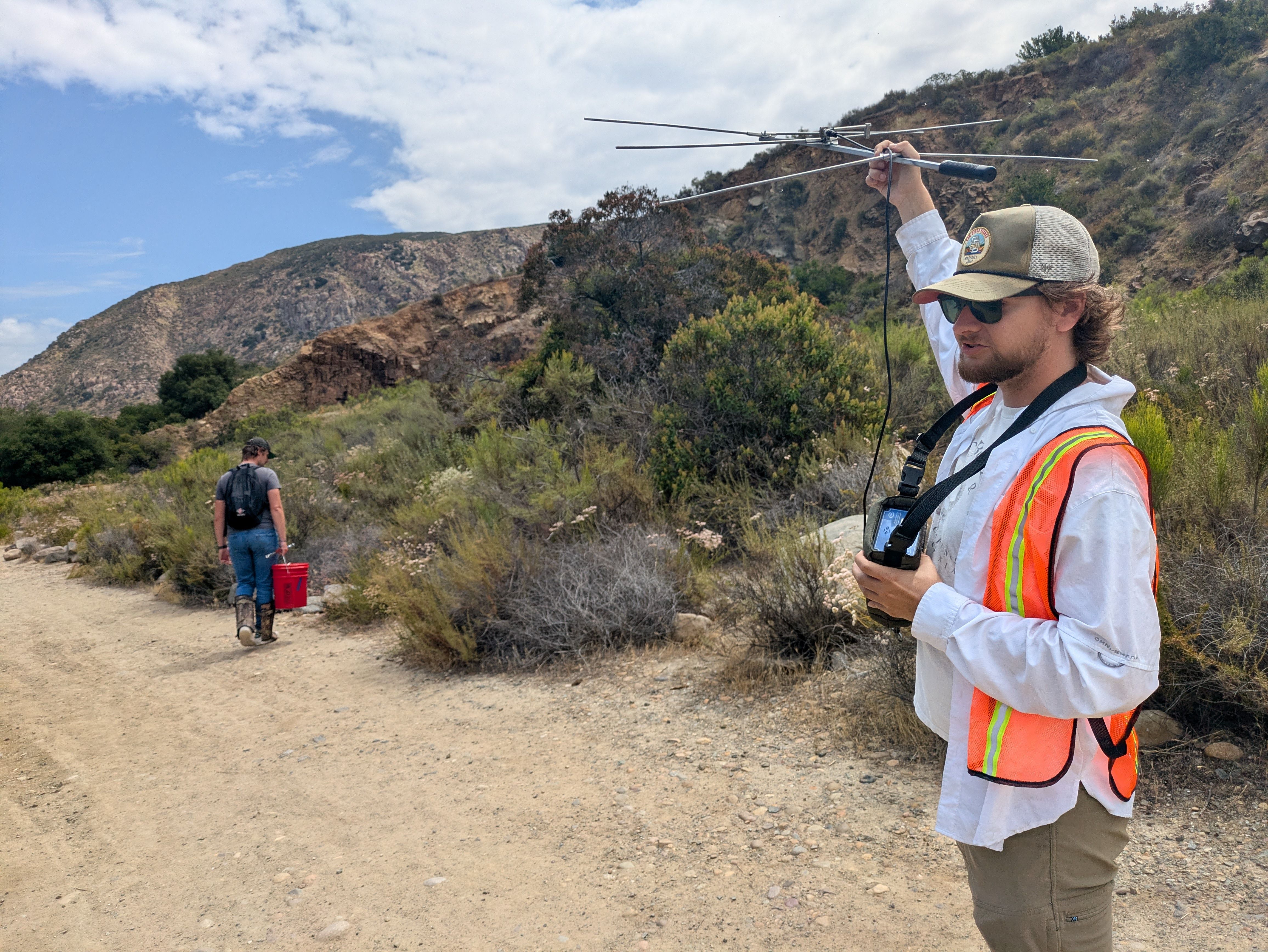 Man wearing a white shirt, orange reflective safety vest, beige cap and sunglasses holds a large multi-element antenna array outdoors on a rocky trail; blue sky and hillside behind.