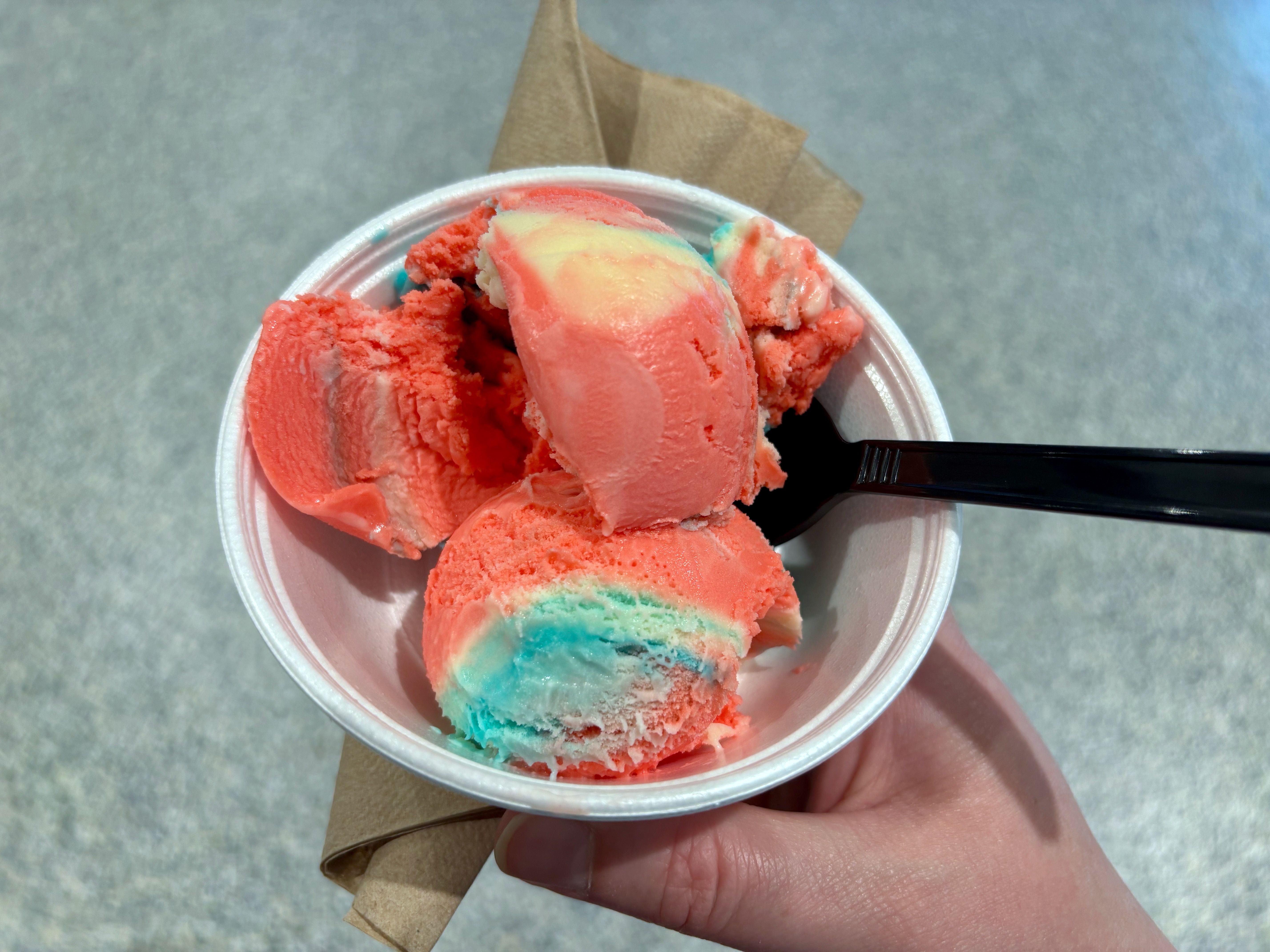 Top-down view of a white cup with scoops of pink, white, and turquoise rainbow ice cream; a hand holds it over a brown napkin with a black plastic spoon nearby.