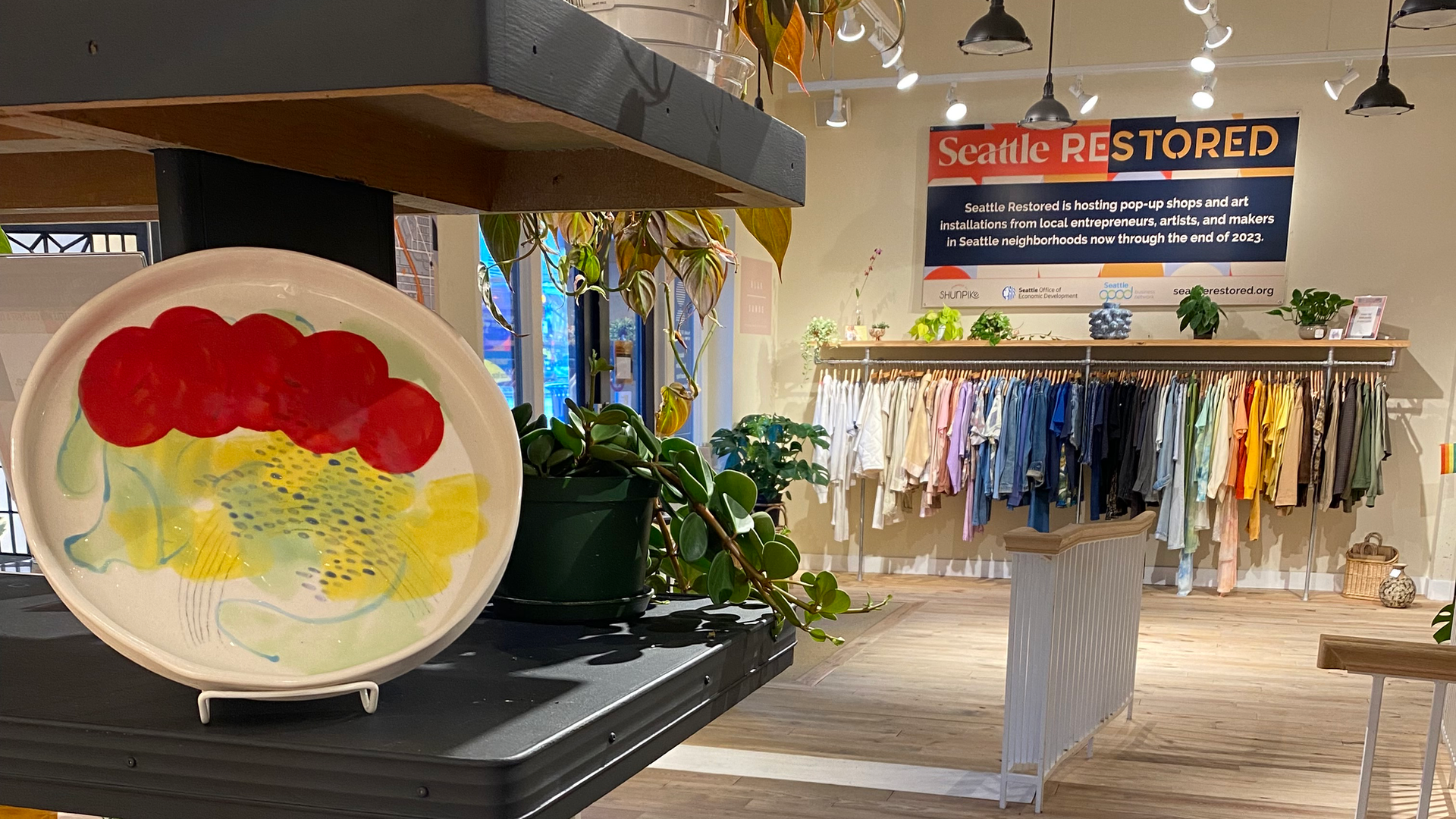 Shelves with an art item on them and a rack of clothes in the background under a sign that says "Seattle Restored." 