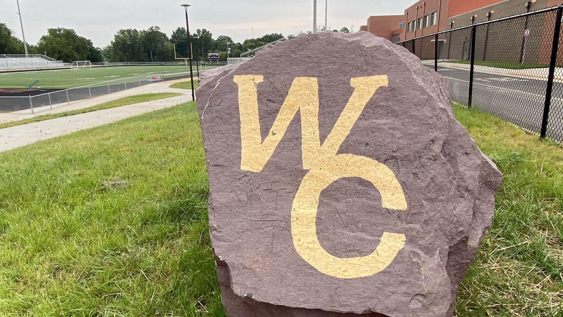 A large purple-gray rock on a grassy patch, painted with big pale-yellow letters "WC". In the background, chain-link fencing, a sports field with goalposts, and a brick building.
