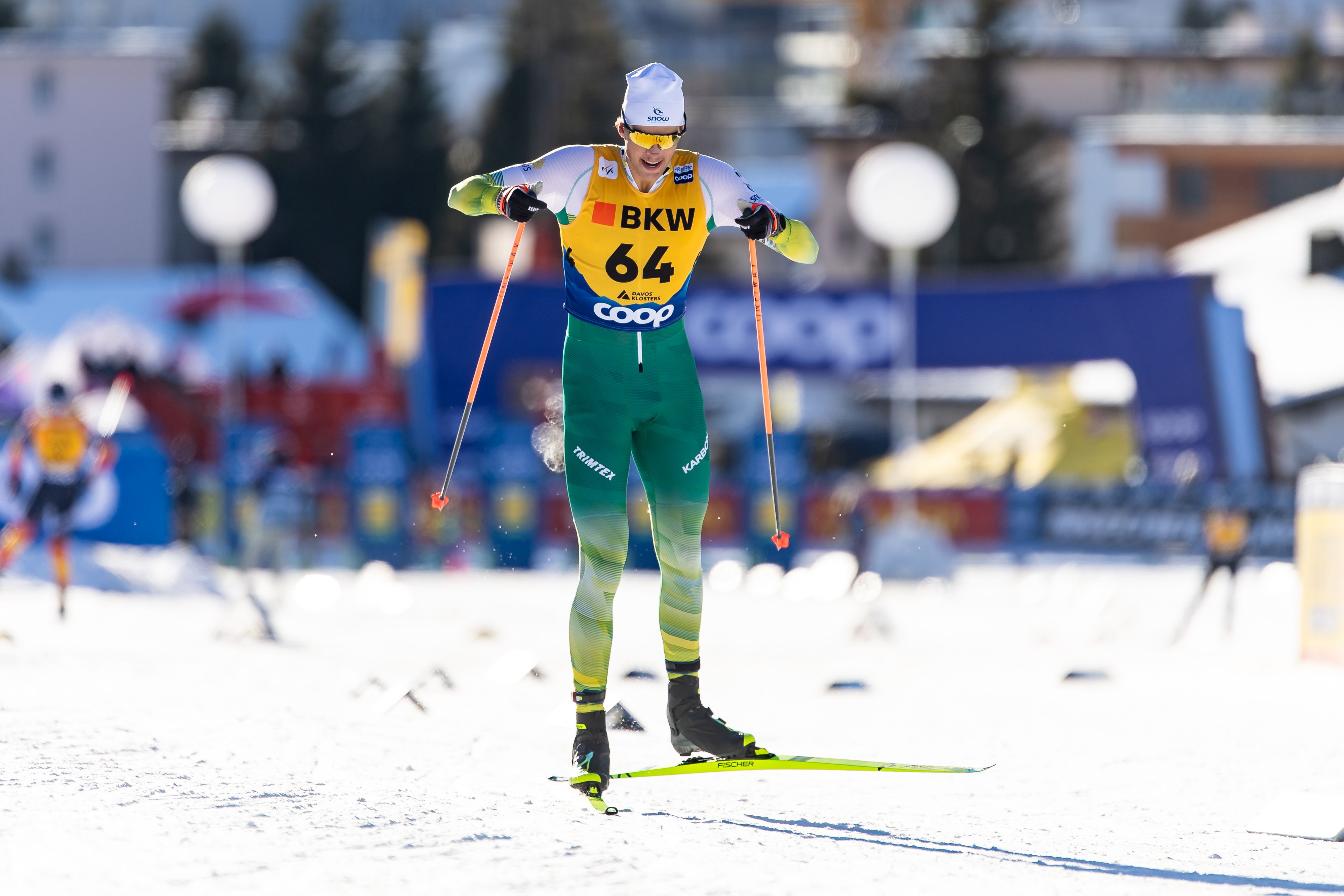Cross-country skier wearing green and white suit, yellow bib number 64, skiing on snow in a race with blurred background and other competitors.