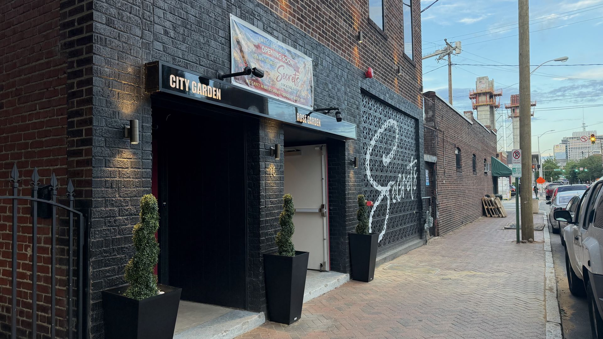 Brick building with black storefronts reading CITY GARDEN and ROSE GARDEN; a banner says Opening Soon. Tall potted evergreens flank the entrance; lattice wall along the side; street with cars under a blue sky.