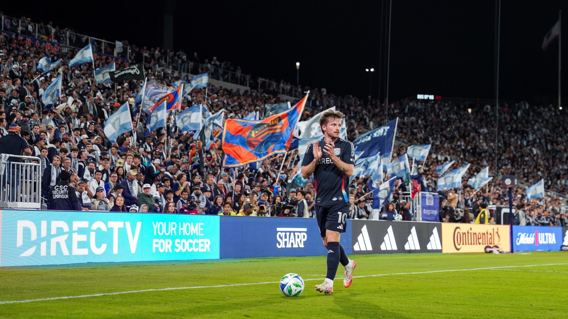 An SDFC player dribbles a ball on the field while clapping with fans cheering behind him.