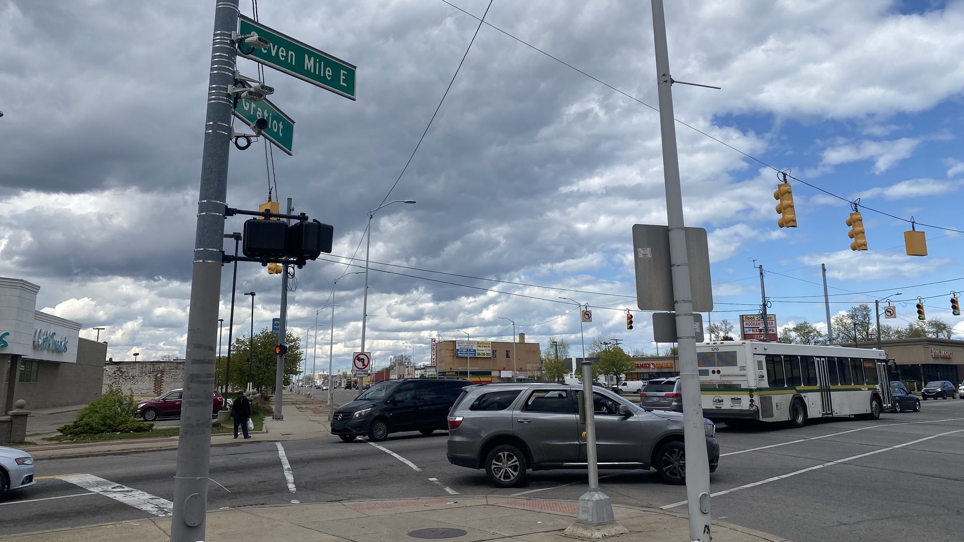 Intersection at Seven Mile E with Gratiot, traffic signals and overhead wires; a city bus and several cars on the street under a cloudy sky with blue patches.