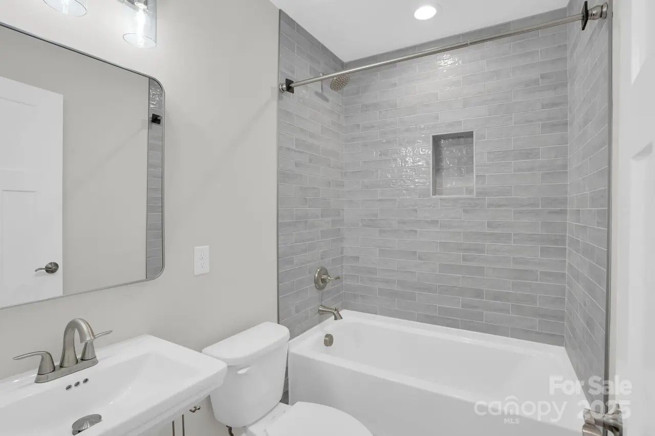 Modern bathroom with white sink, toilet, and bathtub. Gray subway tile surrounds the tub with built-in shelf, silver fixtures, a rectangular mirror, and light gray wall color.