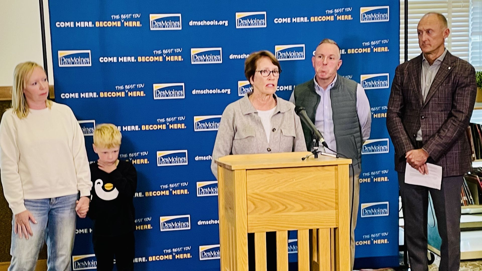 Five people stand in front of a blue Des Moines Public Schools backdrop with the slogan "Come here. Become here." A woman speaks at a wooden podium with a microphone.