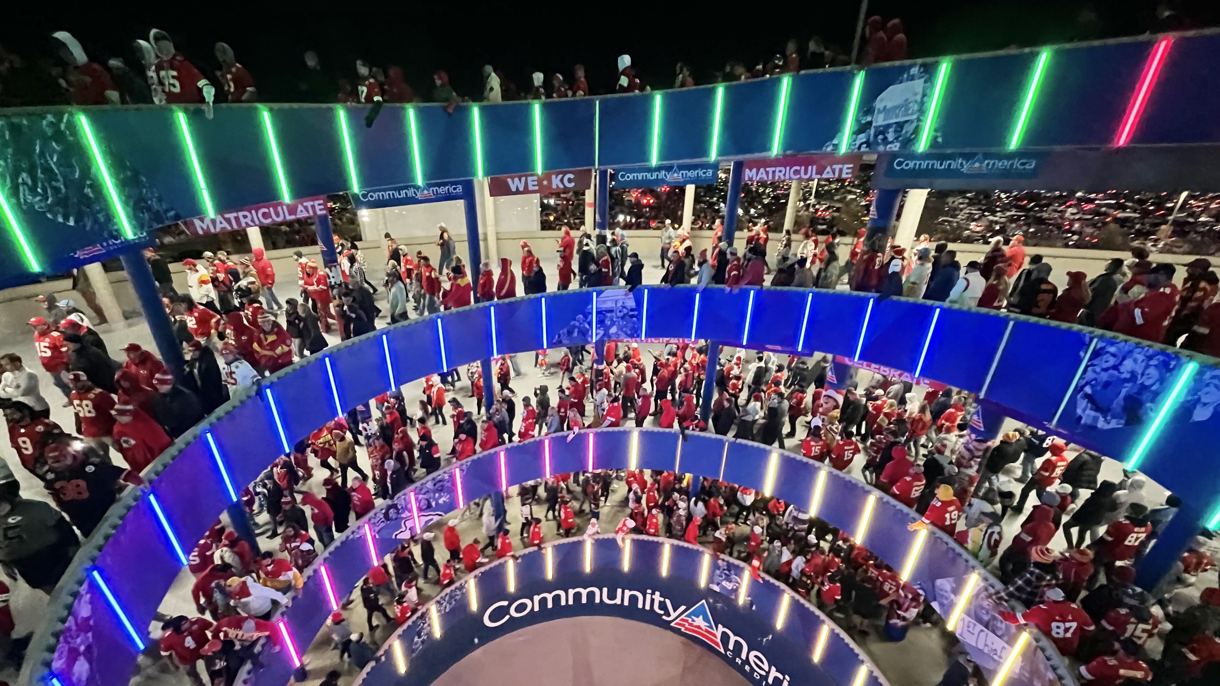 A nighttime view of a circular, multi-level stadium concourse crowded with red-clad fans. Blue railings glow with neon green and pink lights as banners read "MATRICULATE" and "Community America".